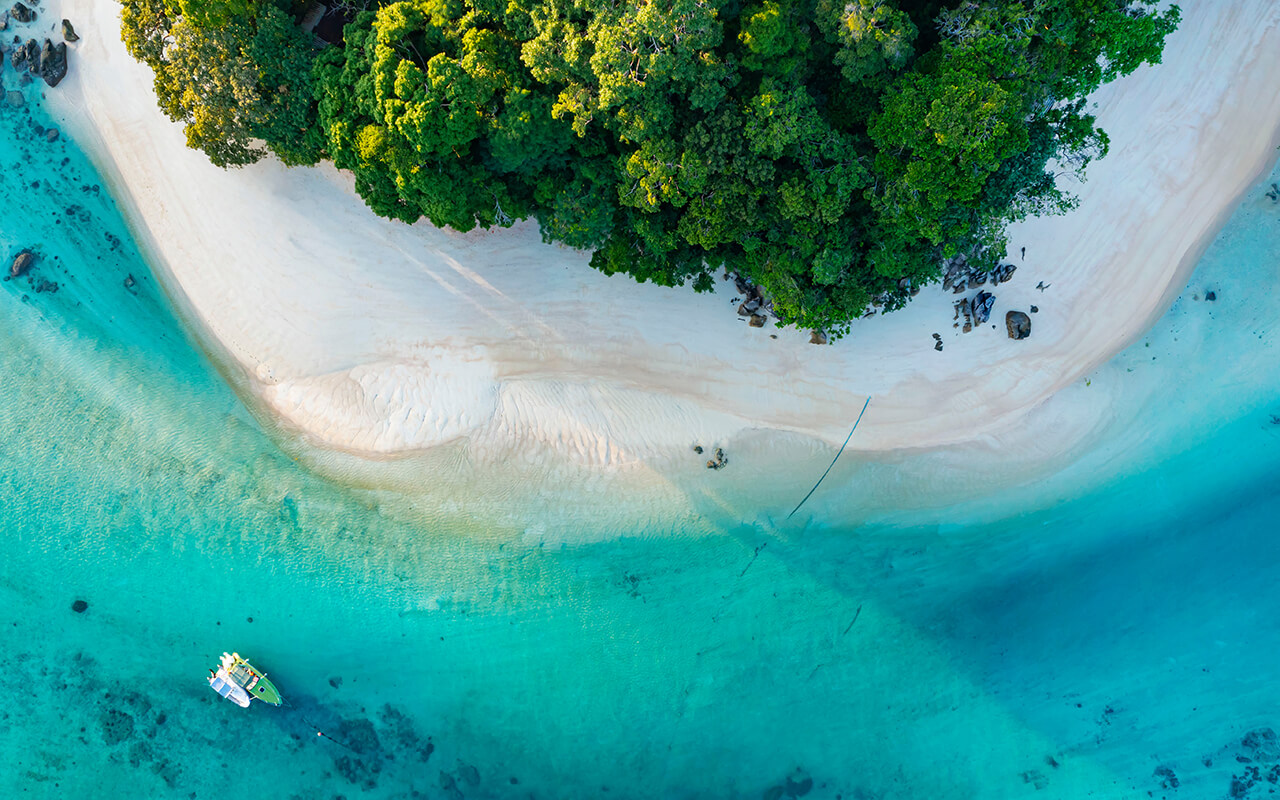 Aerial view of a tropical Pacific island with white sand, turquoise water, and lush green forest.