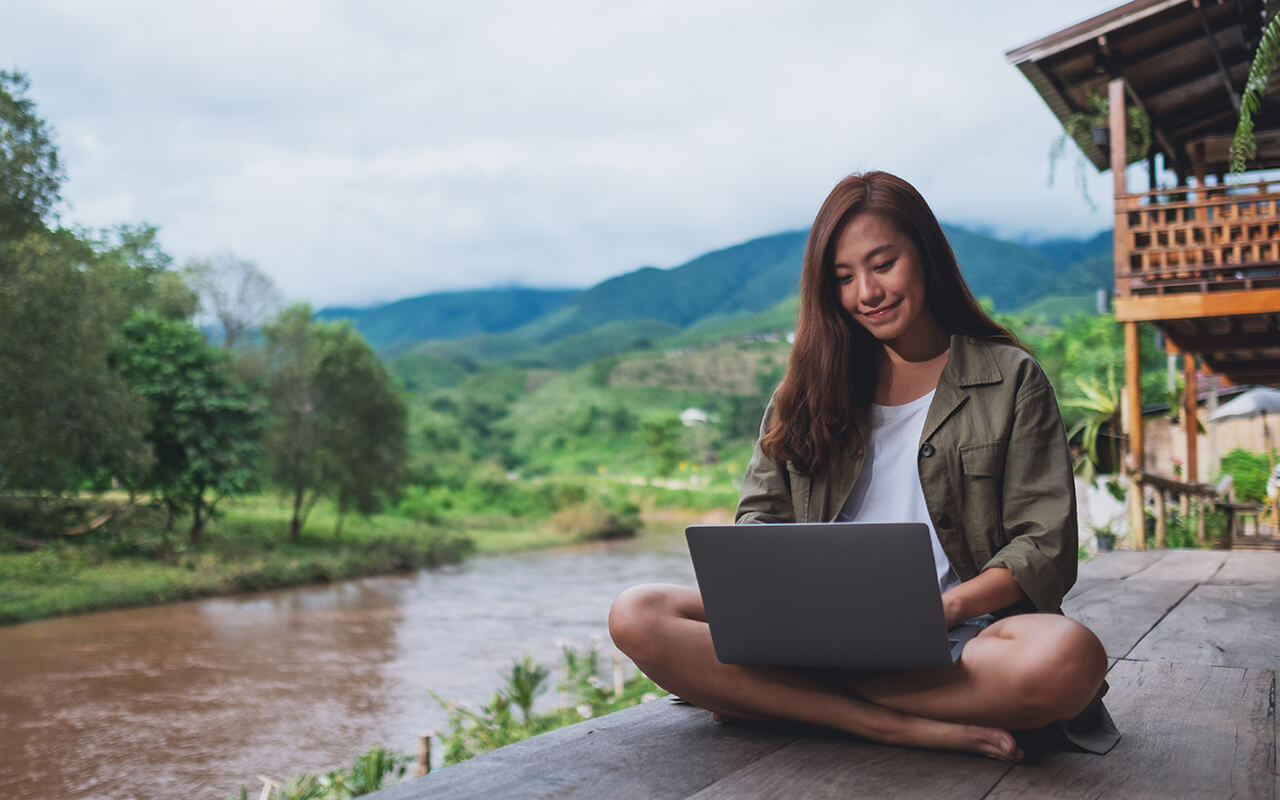 Portrait image of a beautiful asian woman working on laptop computer while sitting by the river with mountains and nature background