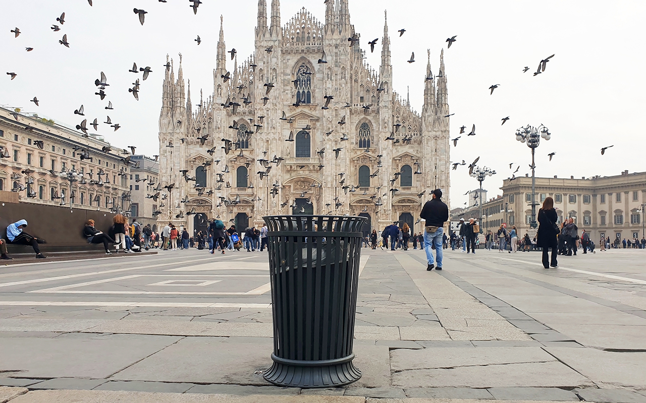 A black metal trash can against the backdrop of the famous Cathedral in Milan with doves flying in the sky.