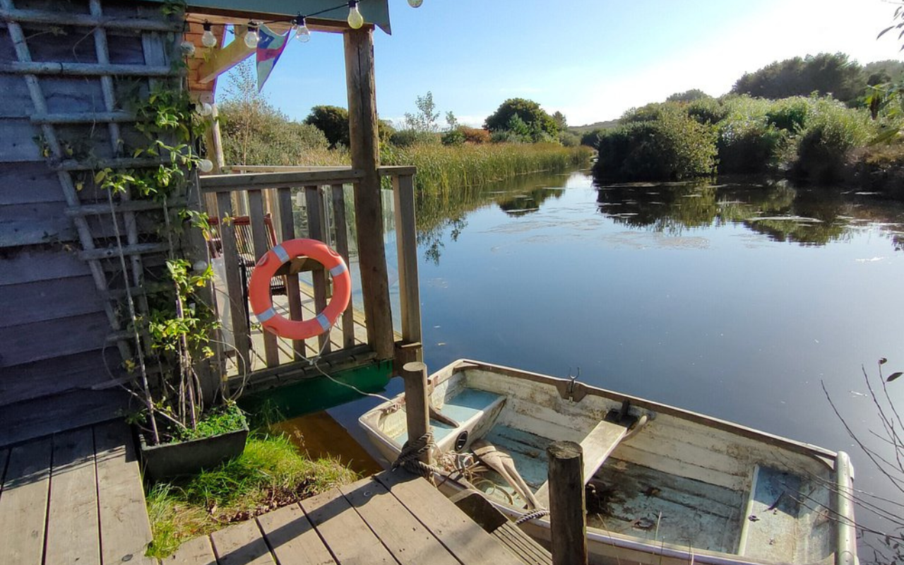 Peaceful view from the dock at The Boat House, Pengelly Retreat in Cornwall, UK, overlooking still waters surrounded by lush greenery and reeds.