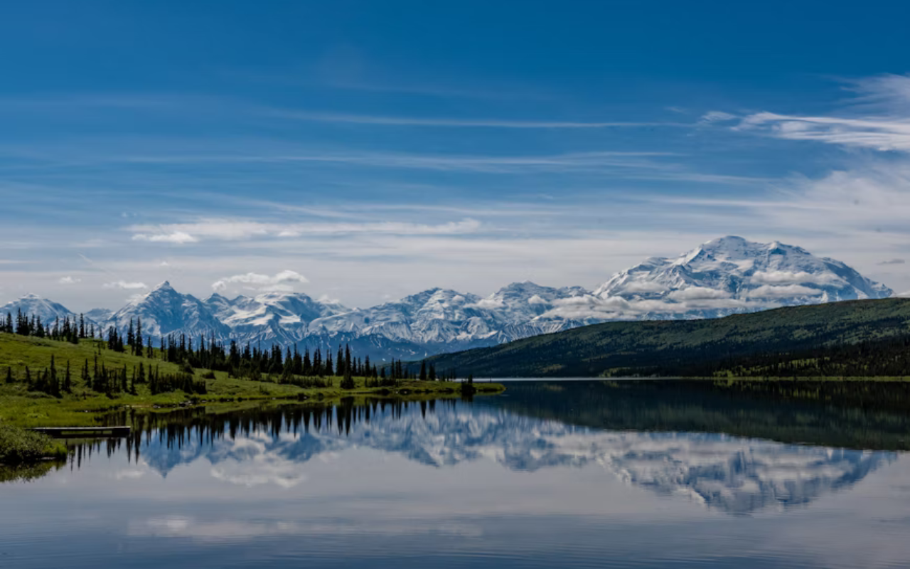 Snow-capped mountains reflect in a calm, clear lake surrounded by lush rolling hills in Denali National Park.