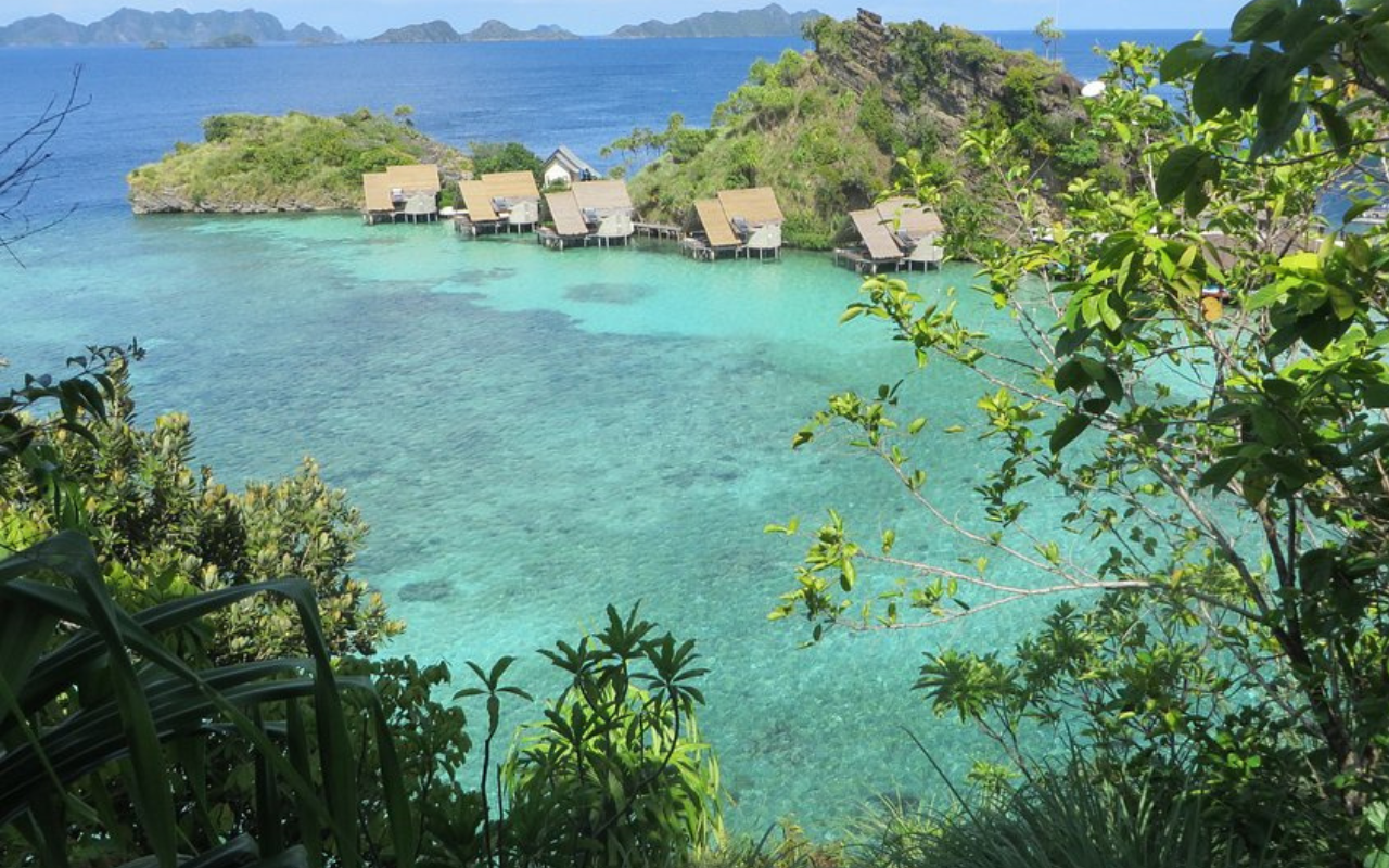 Thatched-roof villas of Misool Eco Resort perched above crystal-clear turquoise waters in Raja Ampat, Indonesia, surrounded by vibrant coral reefs and dramatic limestone islets. The remote tropical setting highlights the resort’s eco-luxury design and pristine marine environment.