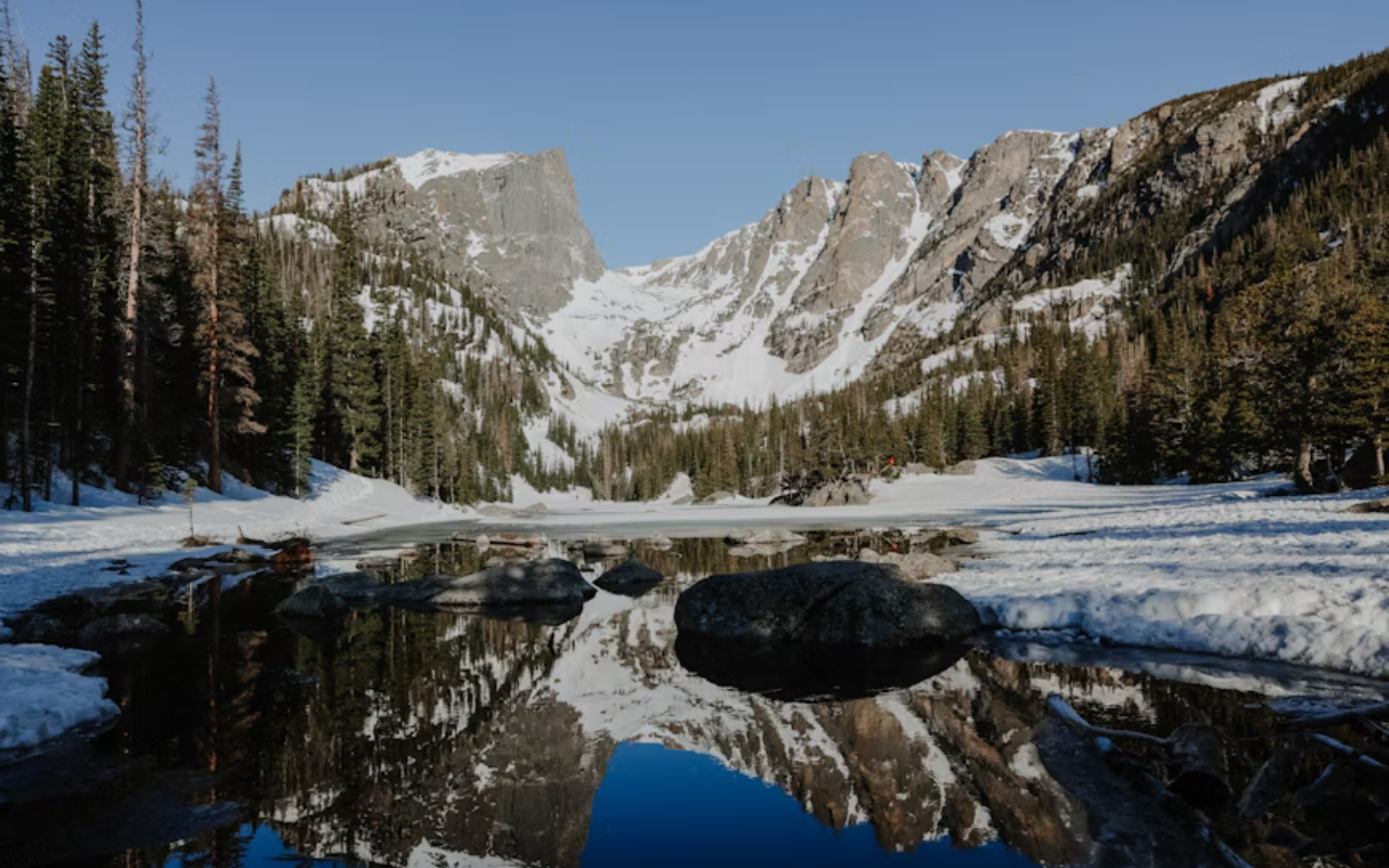 Dense snow coats the ground surrounding a calm river lined by mountains and tall pines in Rocky Mountain National Park.