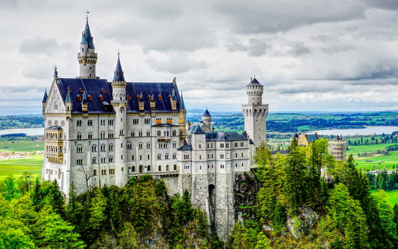 Fairy-tale view of Neuschwanstein Castle perched on a forested hill in the Bavarian Alps, with its white limestone towers and Romanesque Revival architecture surrounded by lush greenery.