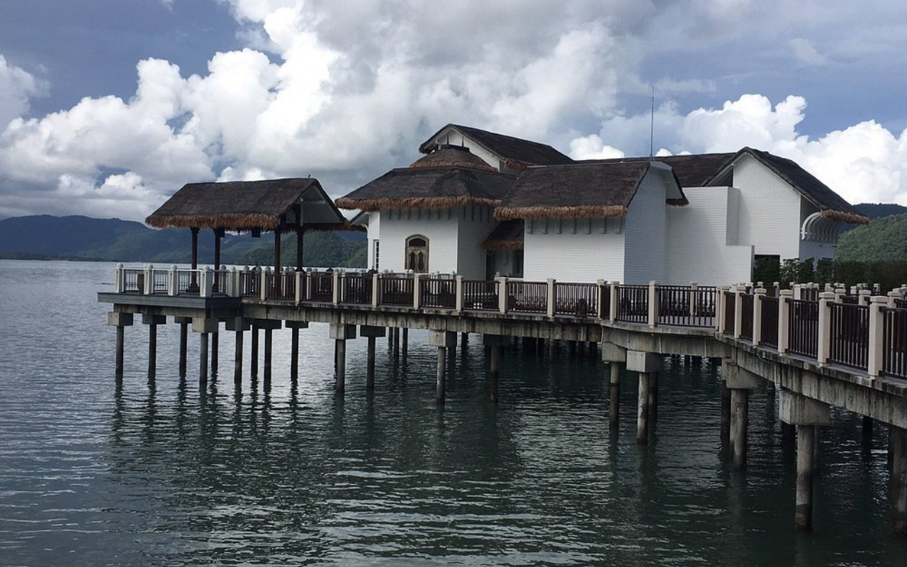 Overwater villas at The St. Regis Langkawi in Malaysia, built on stilts above a calm sea, with white facades, thatched roof accents, and wooden railings. The view captures the peaceful ocean setting under a dramatic sky filled with thick, rolling clouds and distant green hills.
