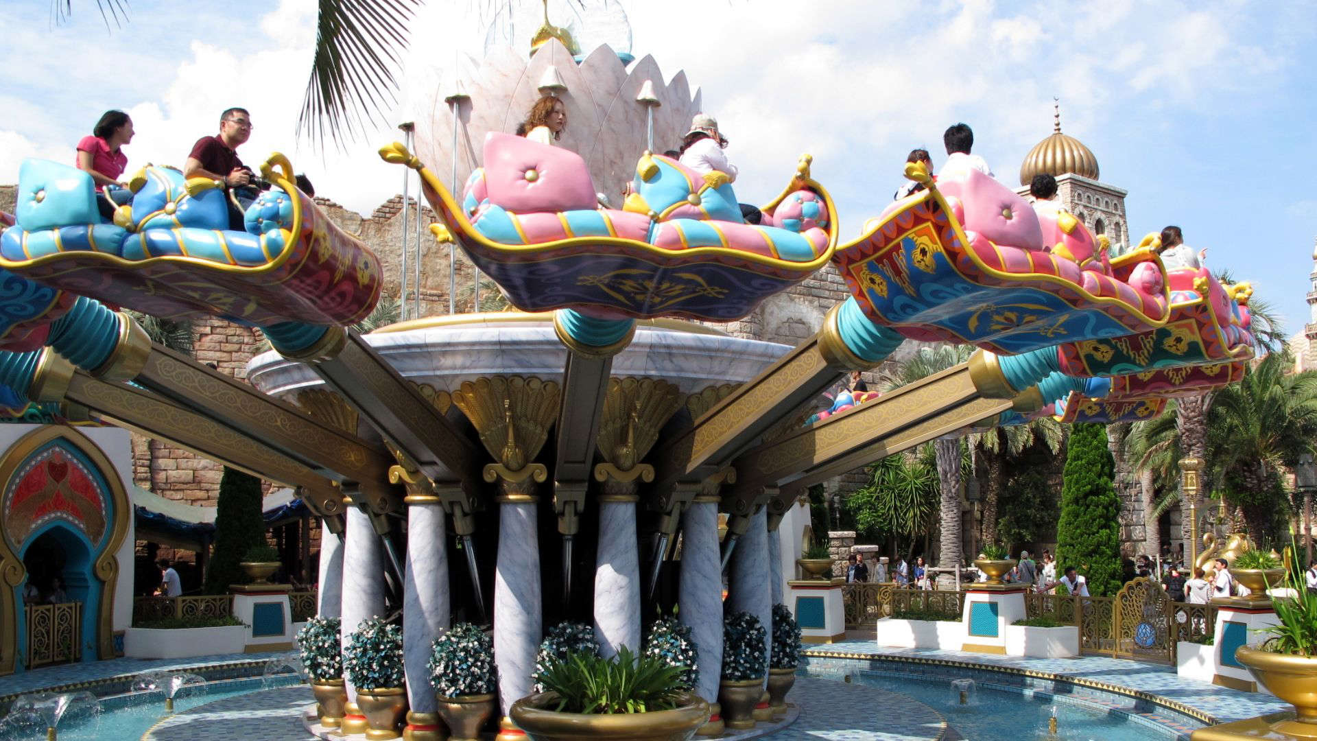 Guests riding colorful flying carpet vehicles on Disney’s The Magic Carpets of Aladdin attraction, soaring gently around a central golden genie lamp.