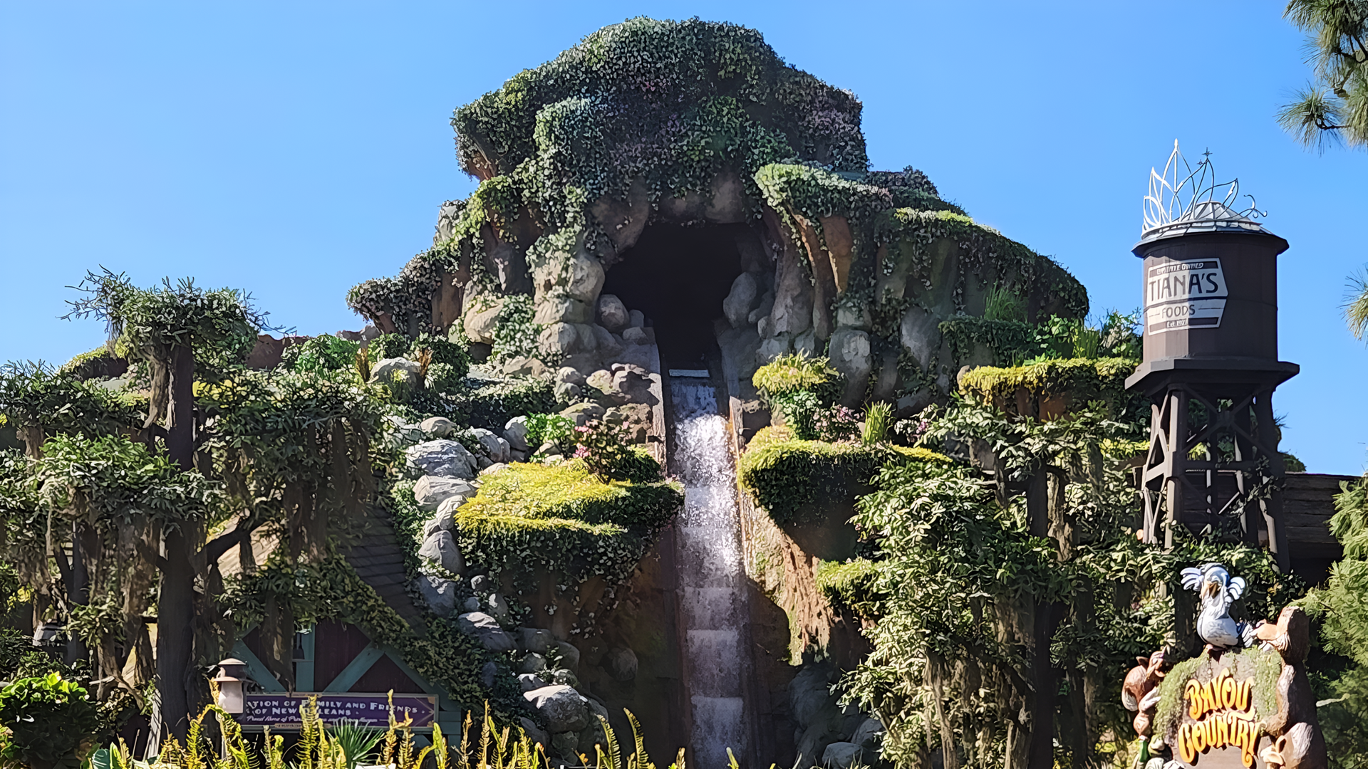 Tiana’s Bayou Adventure, showcasing a lush, green mountainside with a cascading waterfall and a rustic water tower labeled 'Tiana’s Foods.'