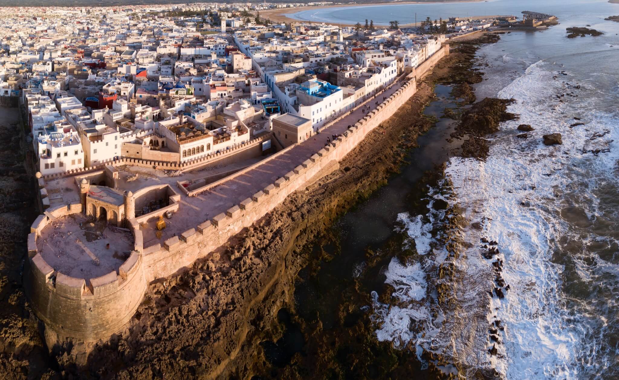 Sunset aerial of Essaouira’s fortified walls and medina along Morocco’s Atlantic coast, showing Skala de la Kasbah and crashing ocean waves