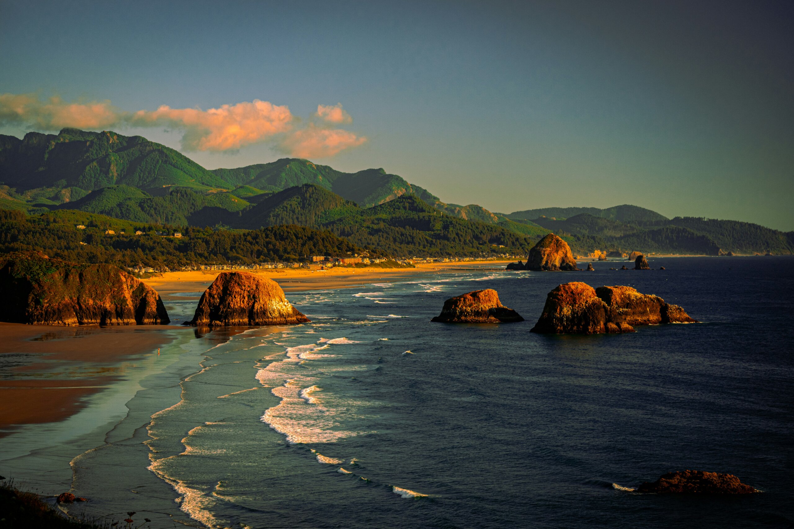 Scenic coastal view of Cannon Beach, Oregon, with sea stacks rising from the Pacific Ocean and lush green forested mountains in the background at golden hour.