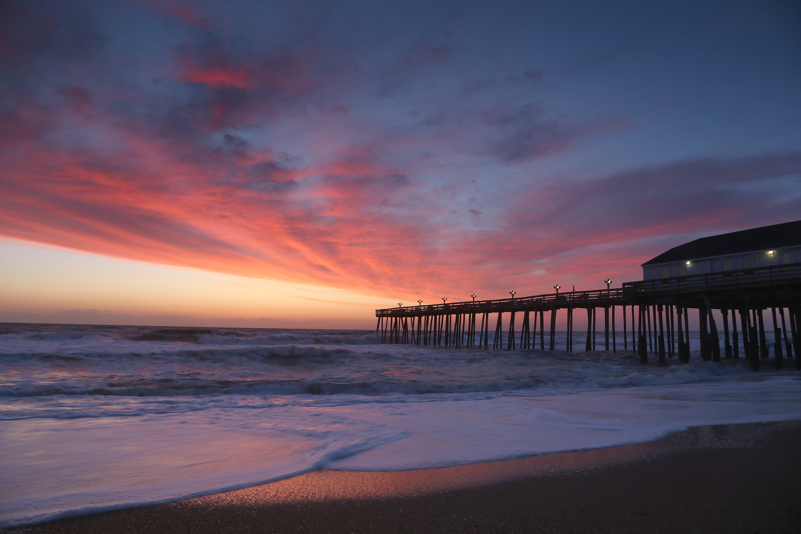 Colorful sunset over the beach and wooden pier in Kitty Hawk, North Carolina, with soft waves washing ashore and warm light reflecting off the ocean.