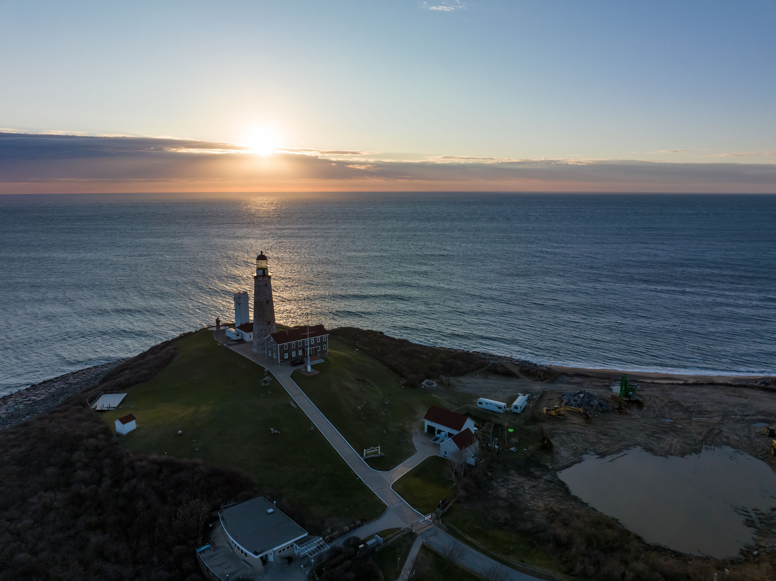 Montauk Lighthouse overlooking the ocean at sunset, with golden light casting a warm glow on the rocky shoreline and waves gently rolling in.
