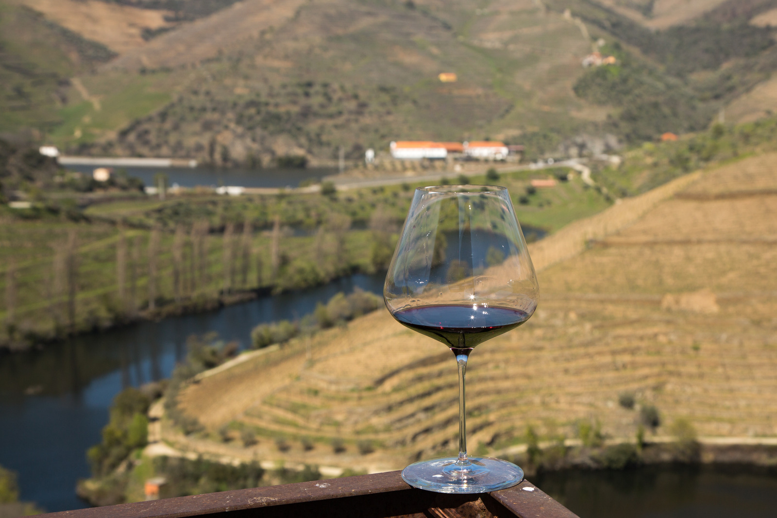 Glass of red wine rests on the railing of a back porch overlooking a river that winds throughout the mountains.