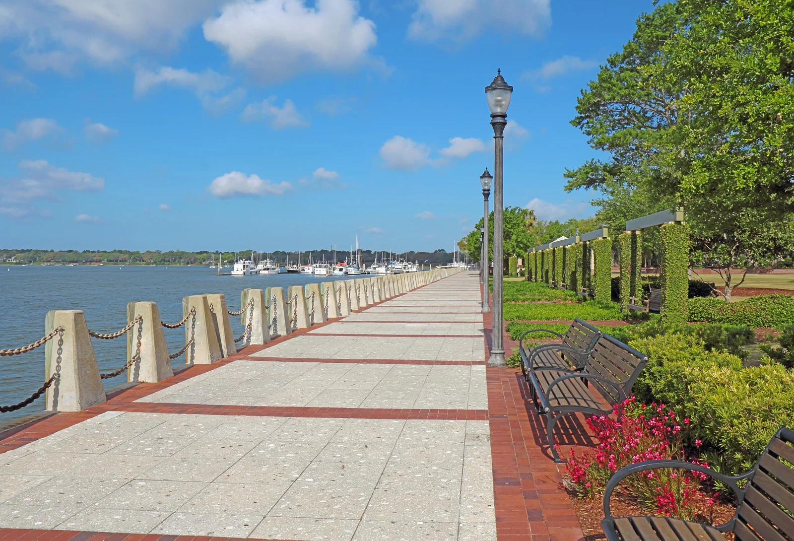 Peaceful waterfront walkway in Beaufort, South Carolina, lined with greenery and benches, overlooking calm waters with boats docked in the background.