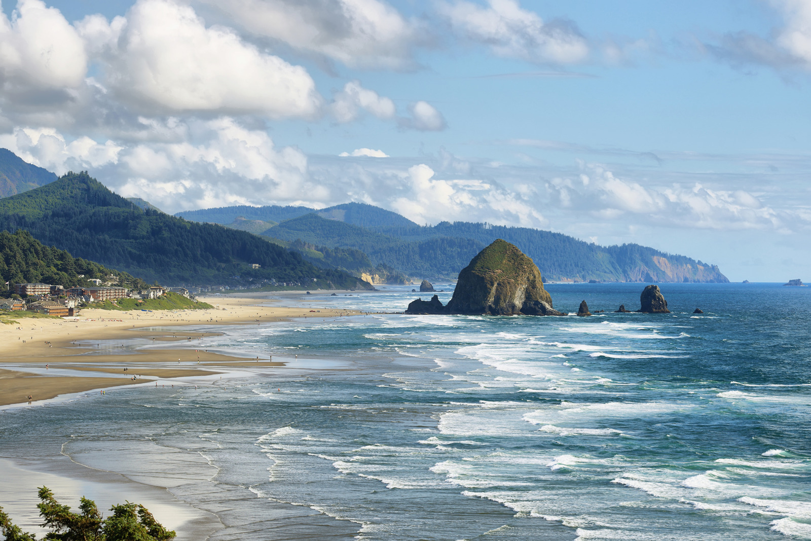 Iconic view of Cannon Beach, Oregon, with Haystack Rock rising from the shoreline, gentle waves rolling in, and misty cliffs in the background beneath a soft coastal sky.