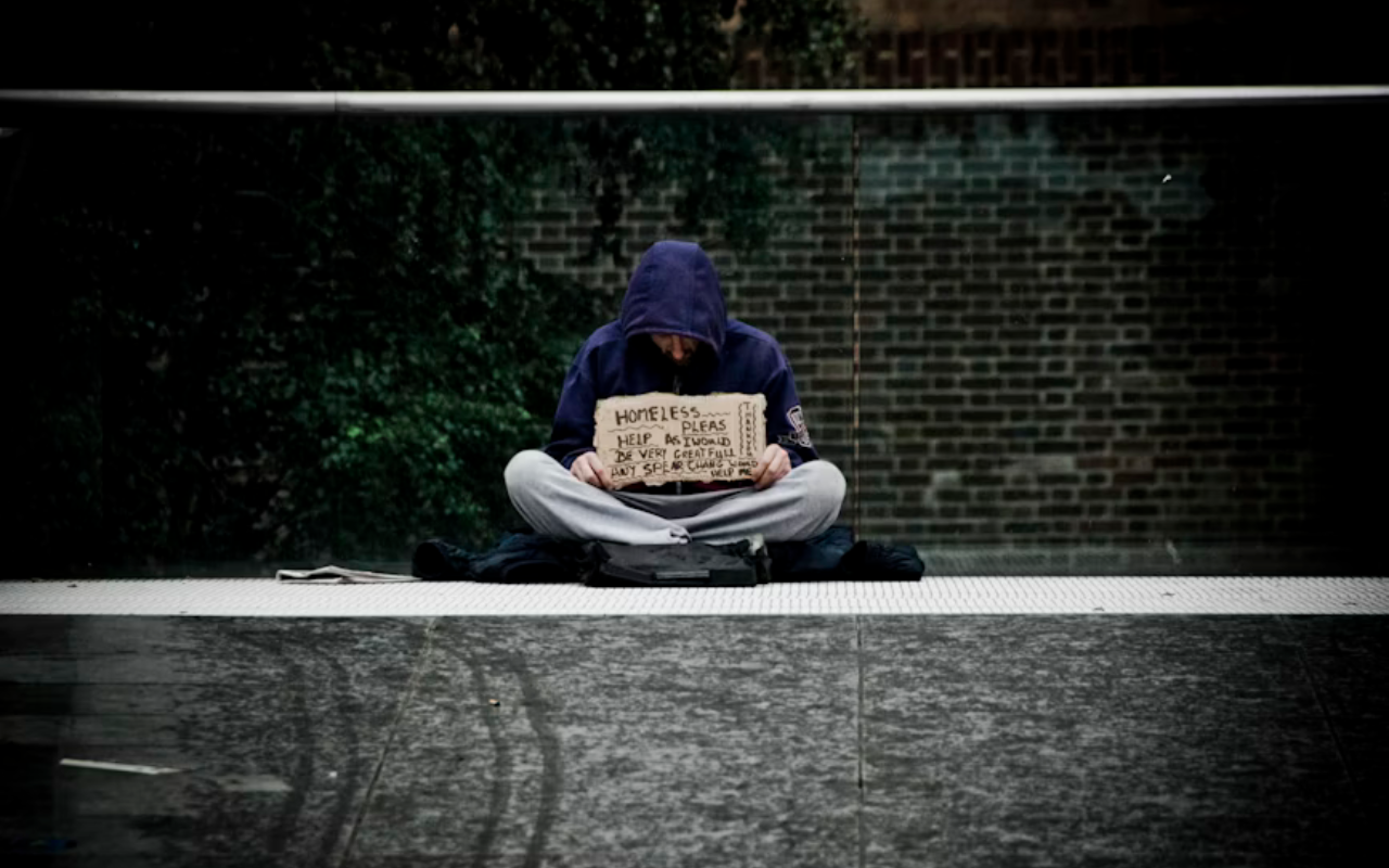 Homeless man with head down and a sign in his hands sitting on a sidewalk in front of a brick building