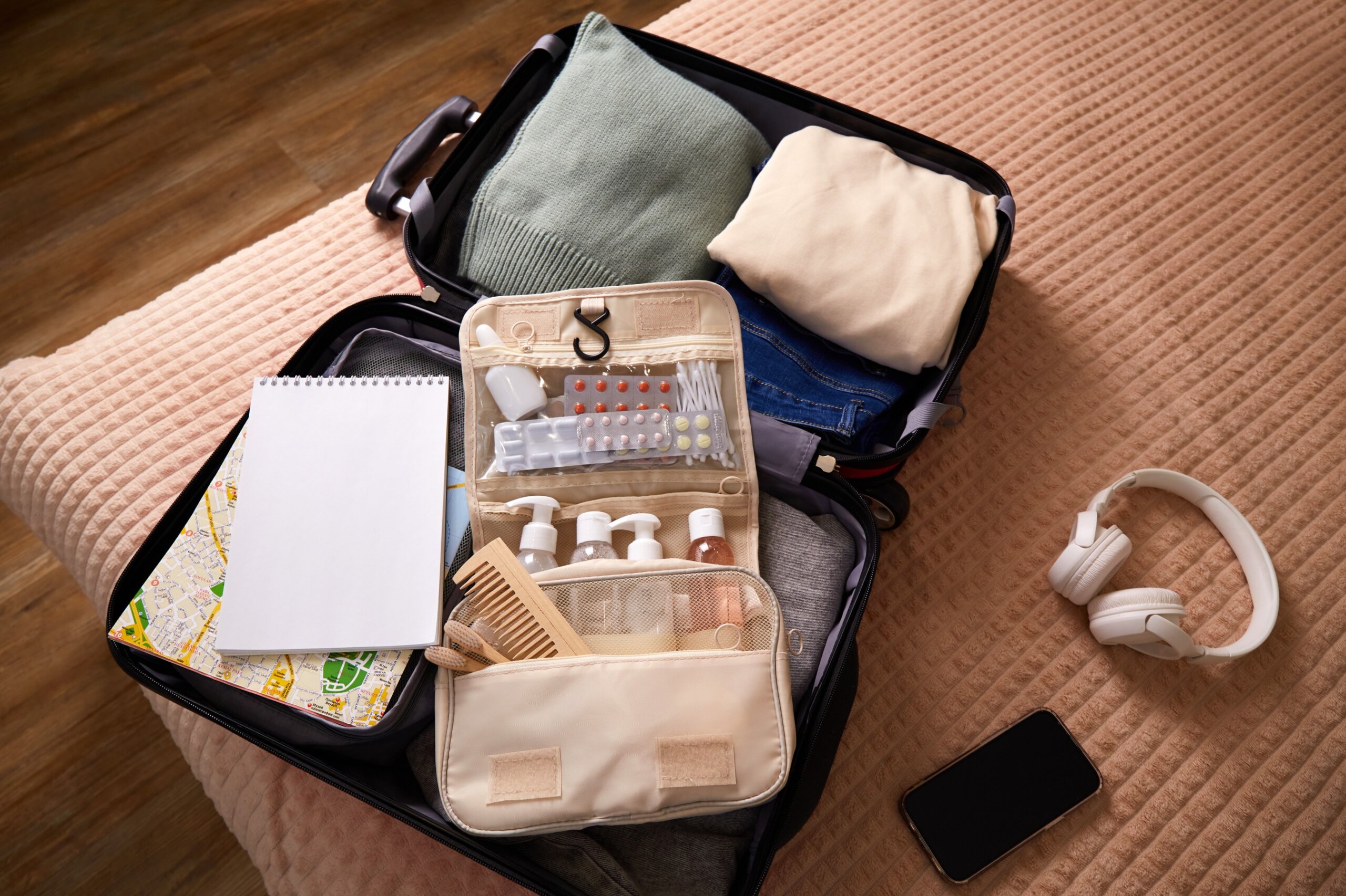A suitcase is open on a bed, containing neatly arranged clothing, toiletries, and travel necessities. A notepad, headphones, and a smartphone are nearby, indicating preparation for a journey