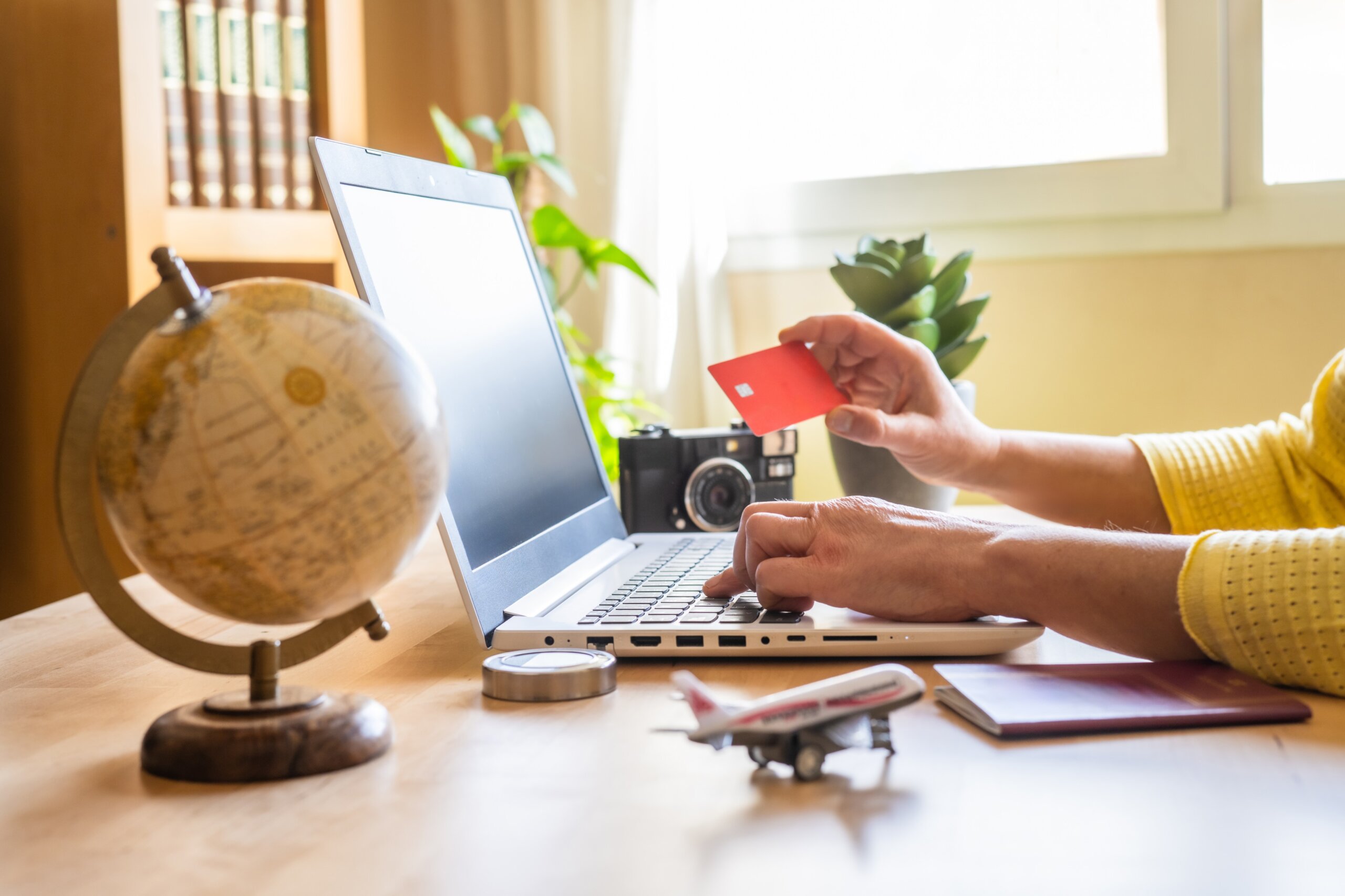 Woman using laptop and credit card to book a trip at home with a globe, passport, toy airplane, and camera on the desk.