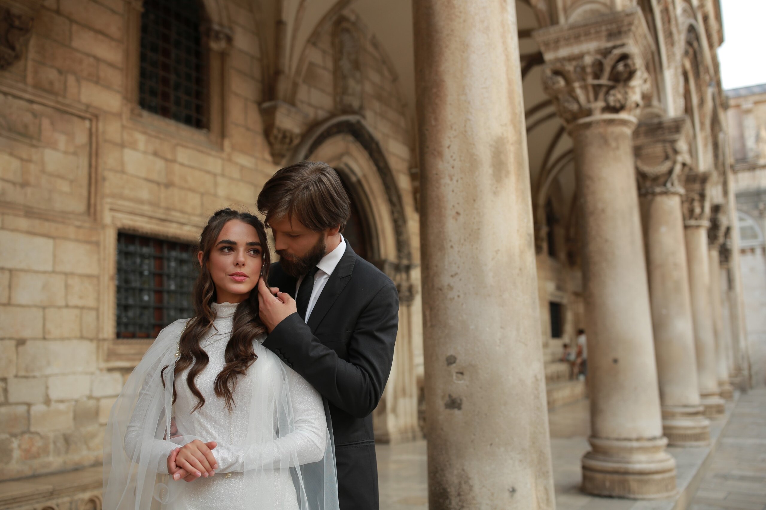 Bride in a long white gown and groom in a suit posing near a historic building in Croatia.