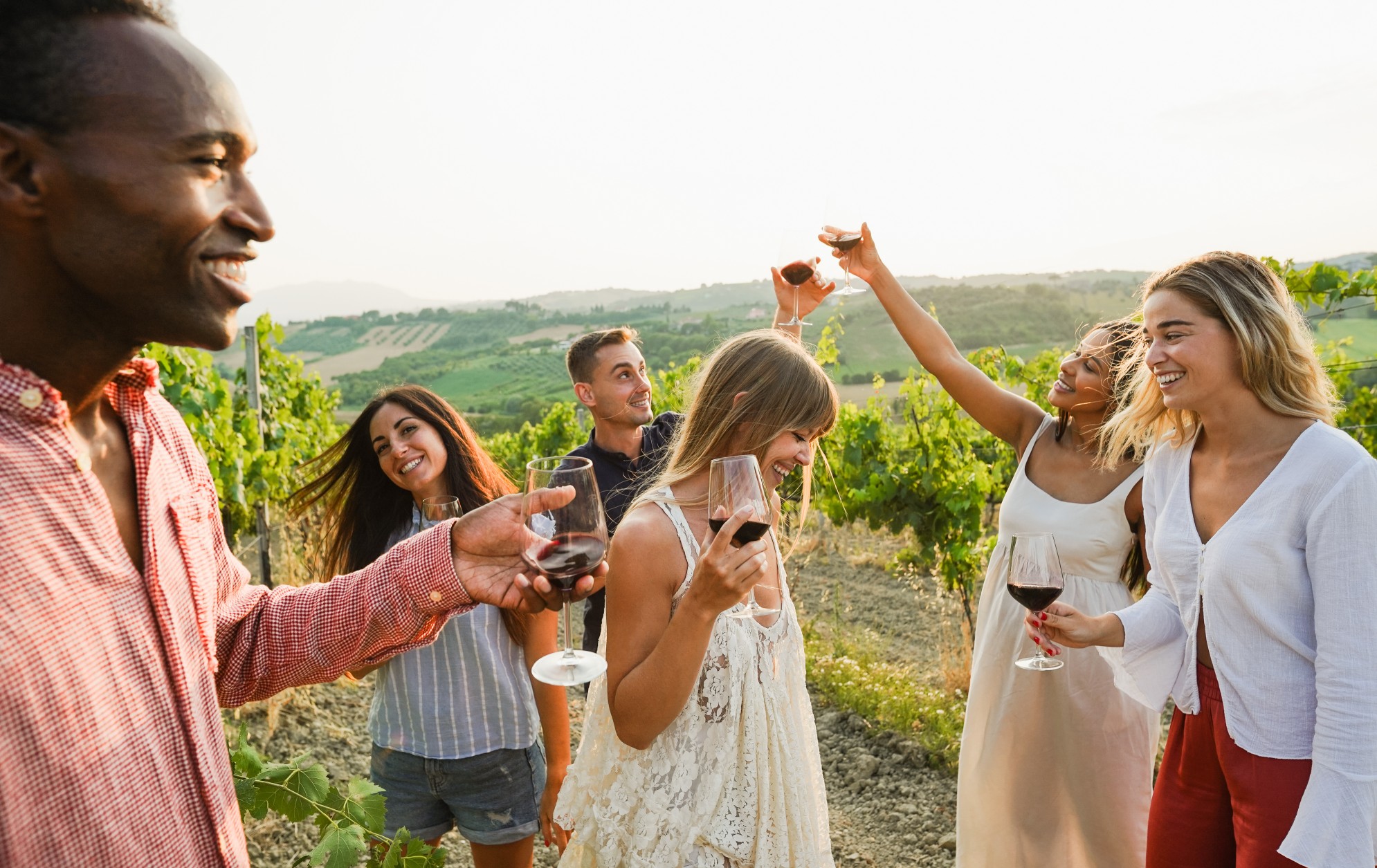 Multiracial friends dancing at summer party inside vineyards - Happy people having fun together drinking red wine at countryside resort - Travel and tasting event concept 