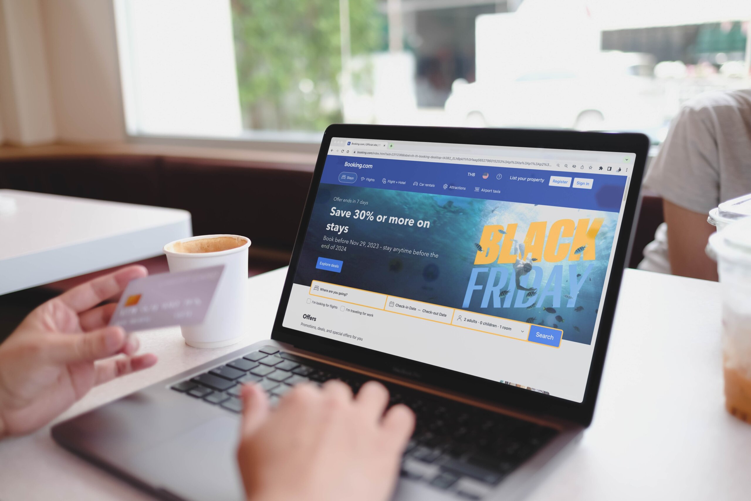 Bangkok, Thailand. May 14, 2025; Person booking hotel on Booking.com website via laptop during Black Friday sale, holding credit card with coffee beside computer.