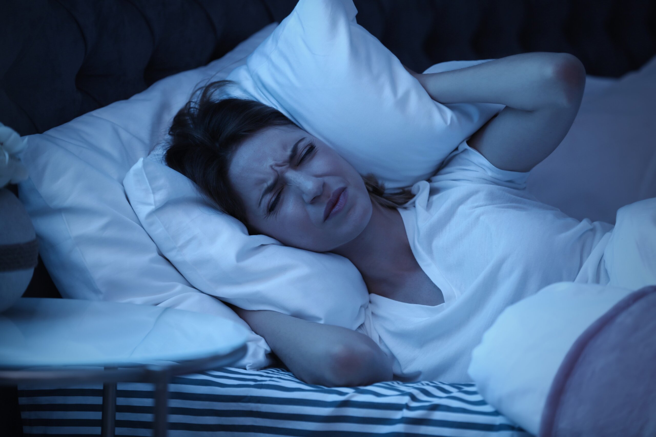 Young woman covering ears with pillow while trying to sleep in bed at night