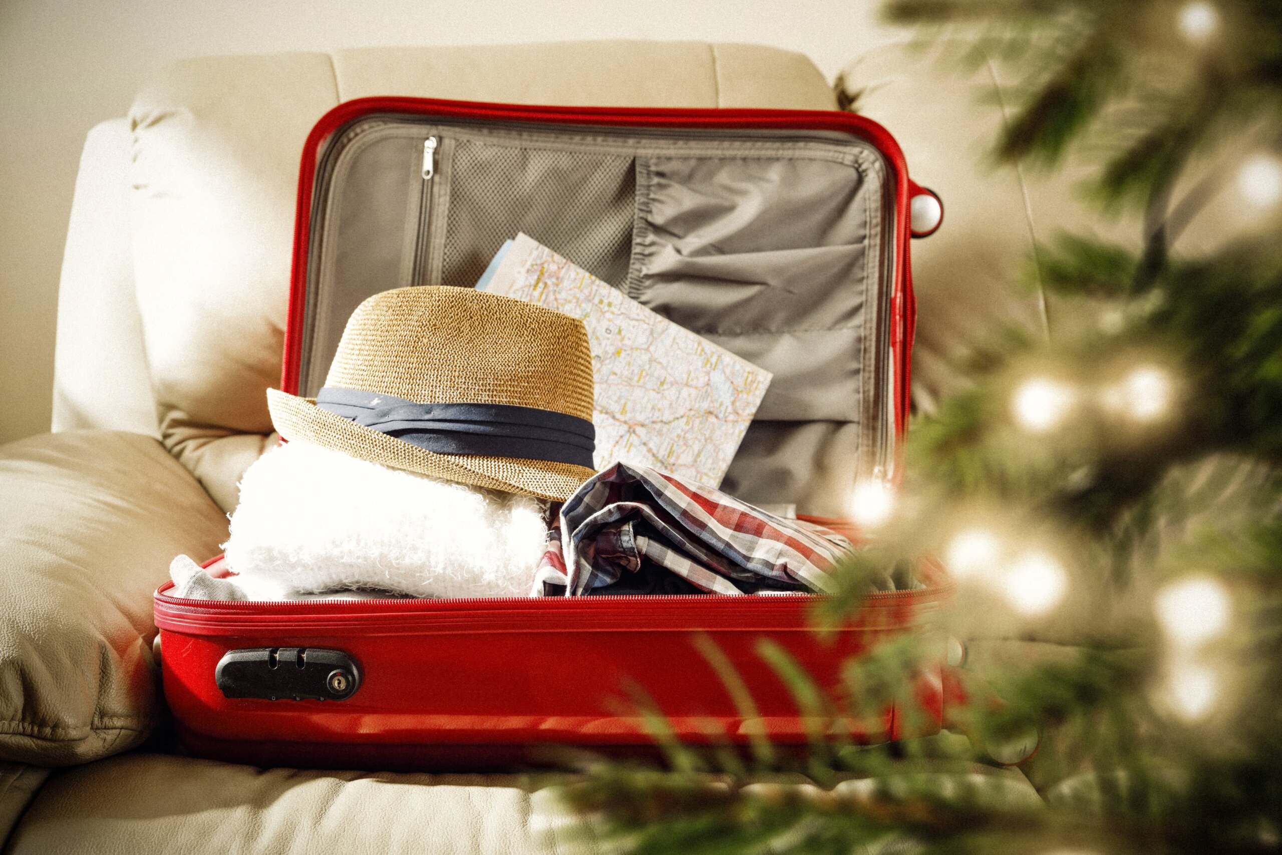 Open suitcase with clothes and a map beside a Christmas tree, representing holiday travel planning and booking flights home for the holidays.