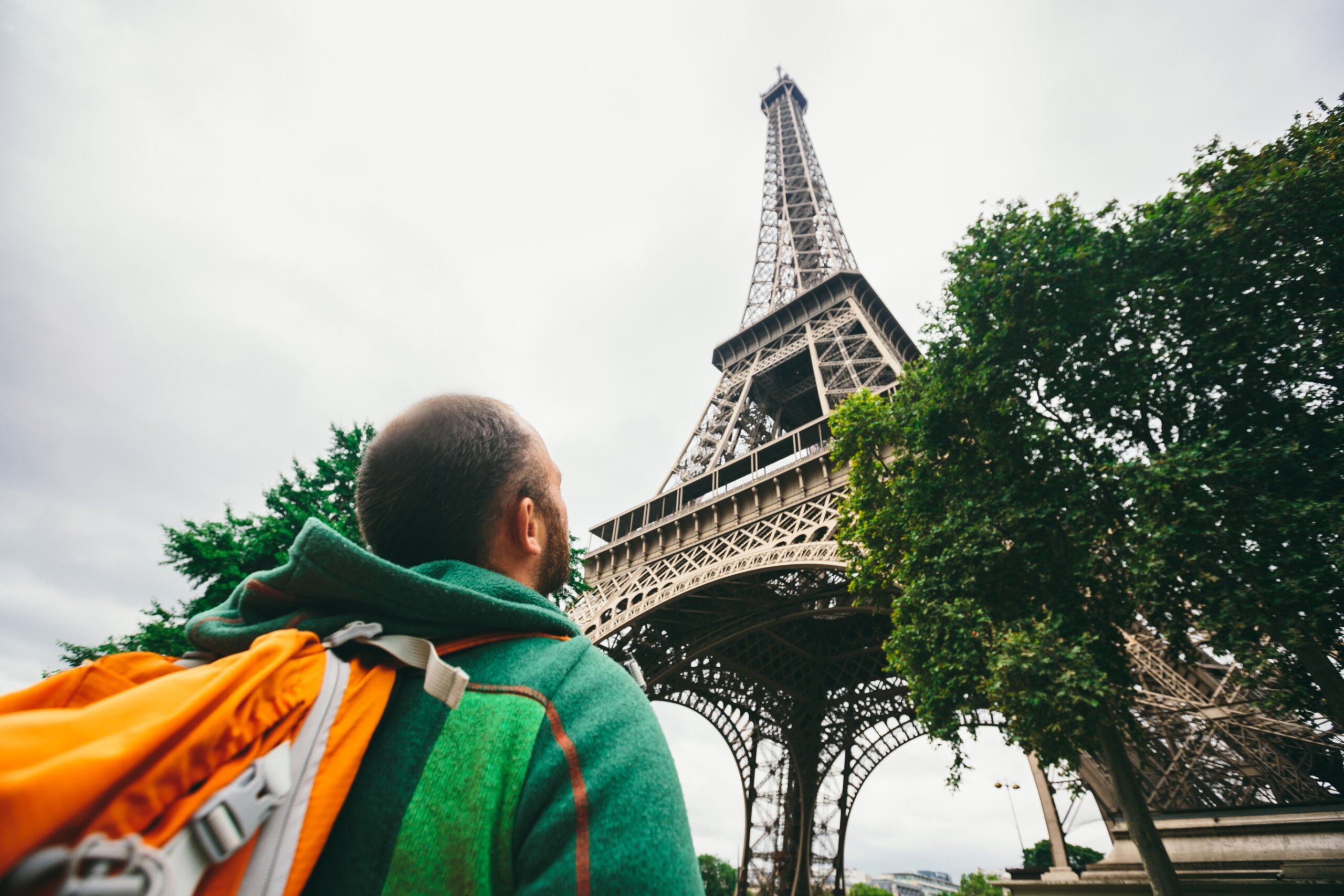 Caucasian male tourist with a backpack looking up at the Eiffel Tower in Paris, France, on a cloudy day.