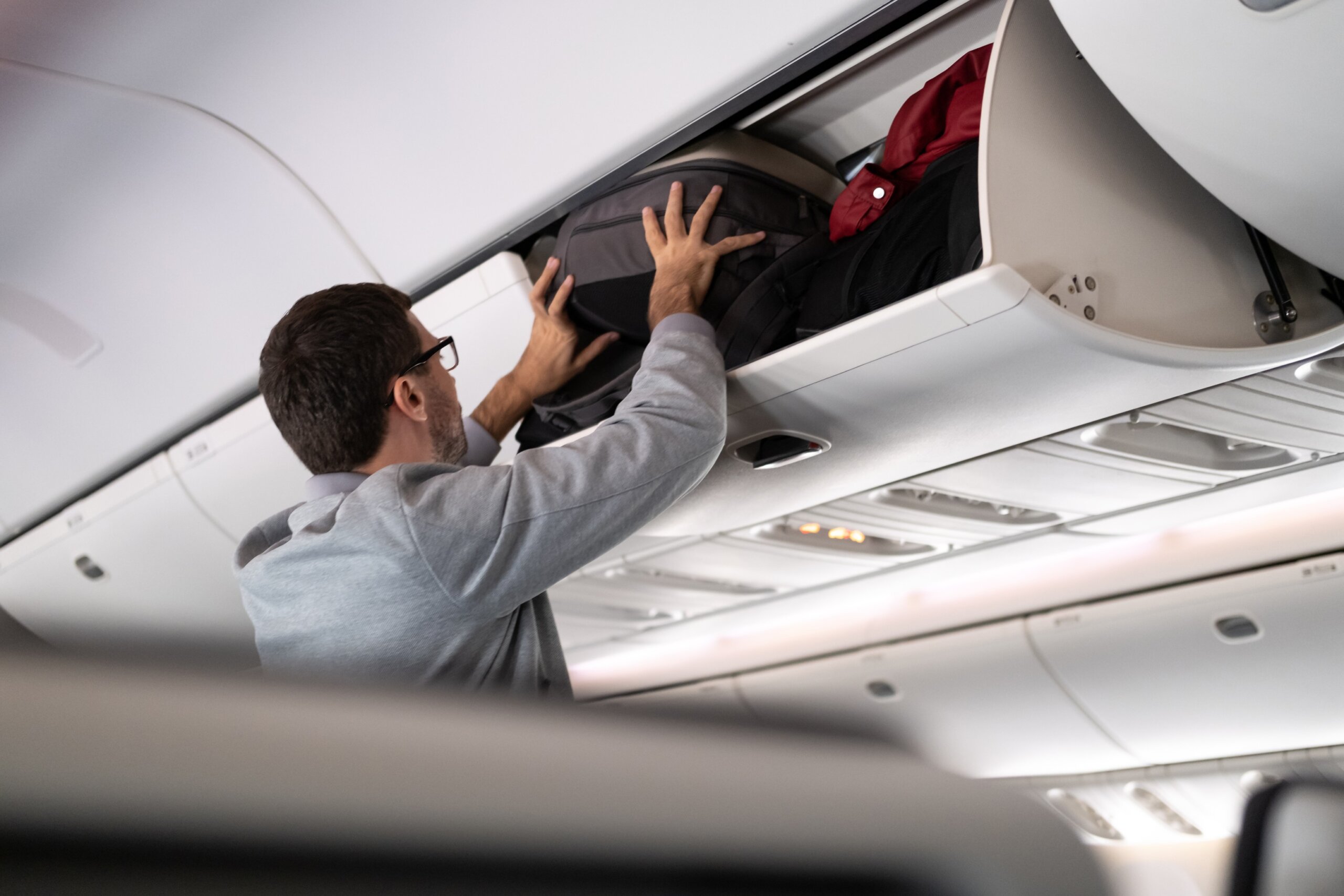 Young man putting luggage into overhead locker on airplane. Traveler placing carry on bag in overhead compartment