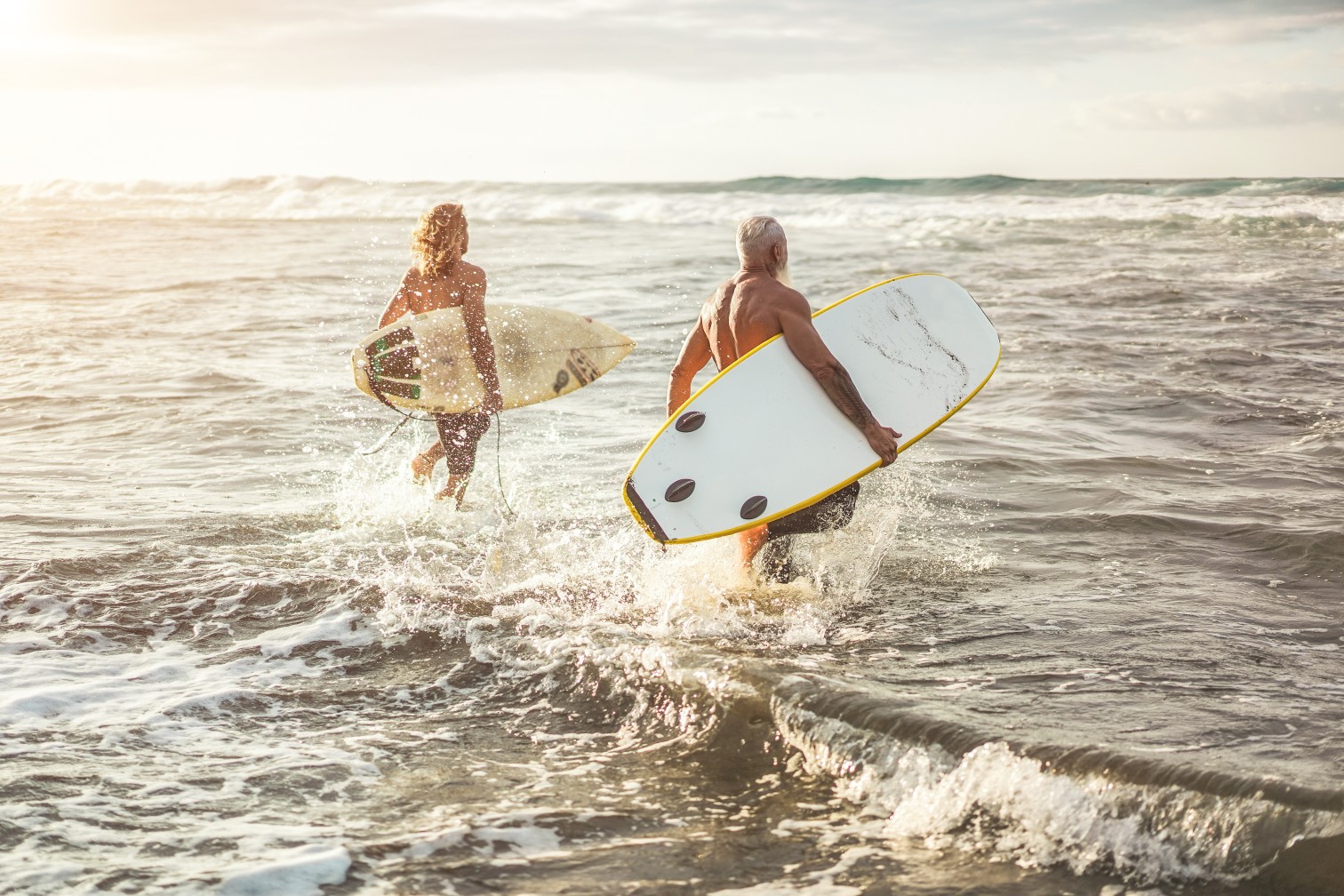 Different age friends running on the beach at sunset for surf training - Family people having fun doing extreme sport - Joyful elderly, travel and healthy lifestyle concept