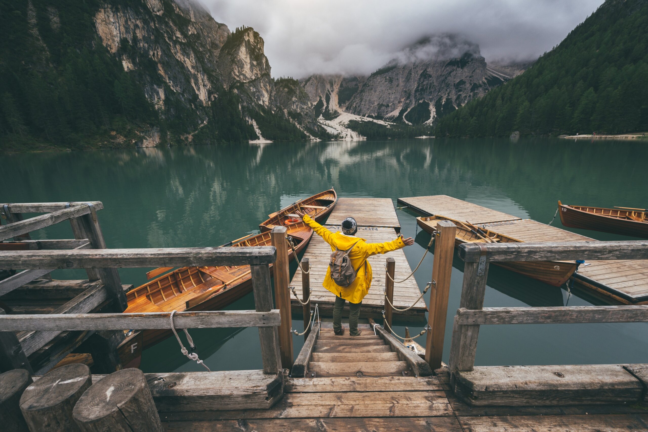 Rear view portrait of a hiker with arms outstretched visiting alpine lake at Braies, Italy. Happy hiker wearing yellow jacket and backpack enjoy the nature landscape at autumn. Wanderlust and travel.