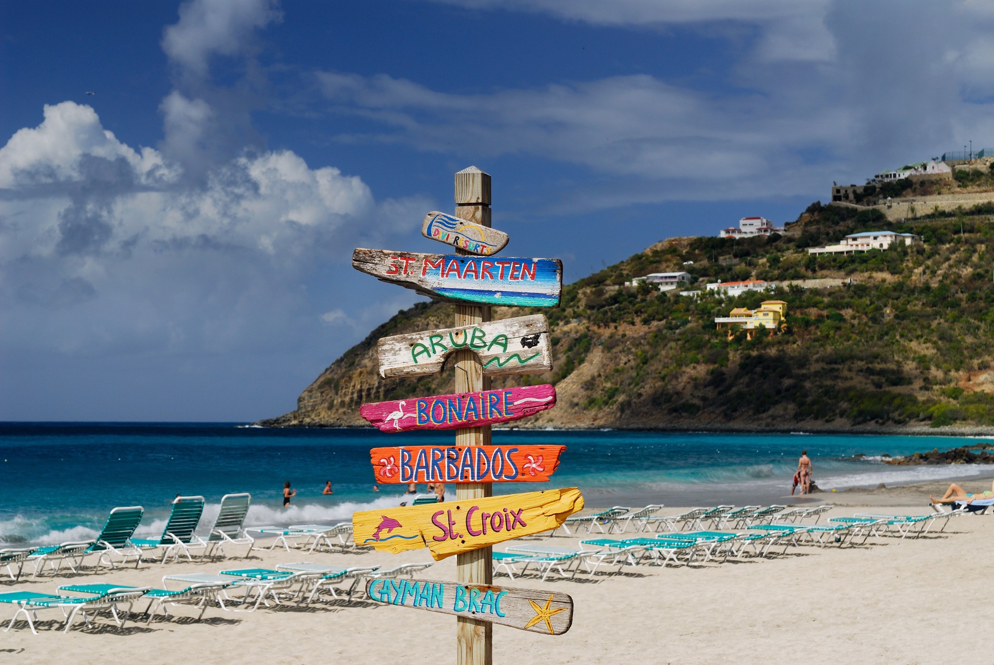 Caribbean beach signpost showing Aruba, Barbados, St. Maarten, and other islands with ocean and lounge chairs in view.