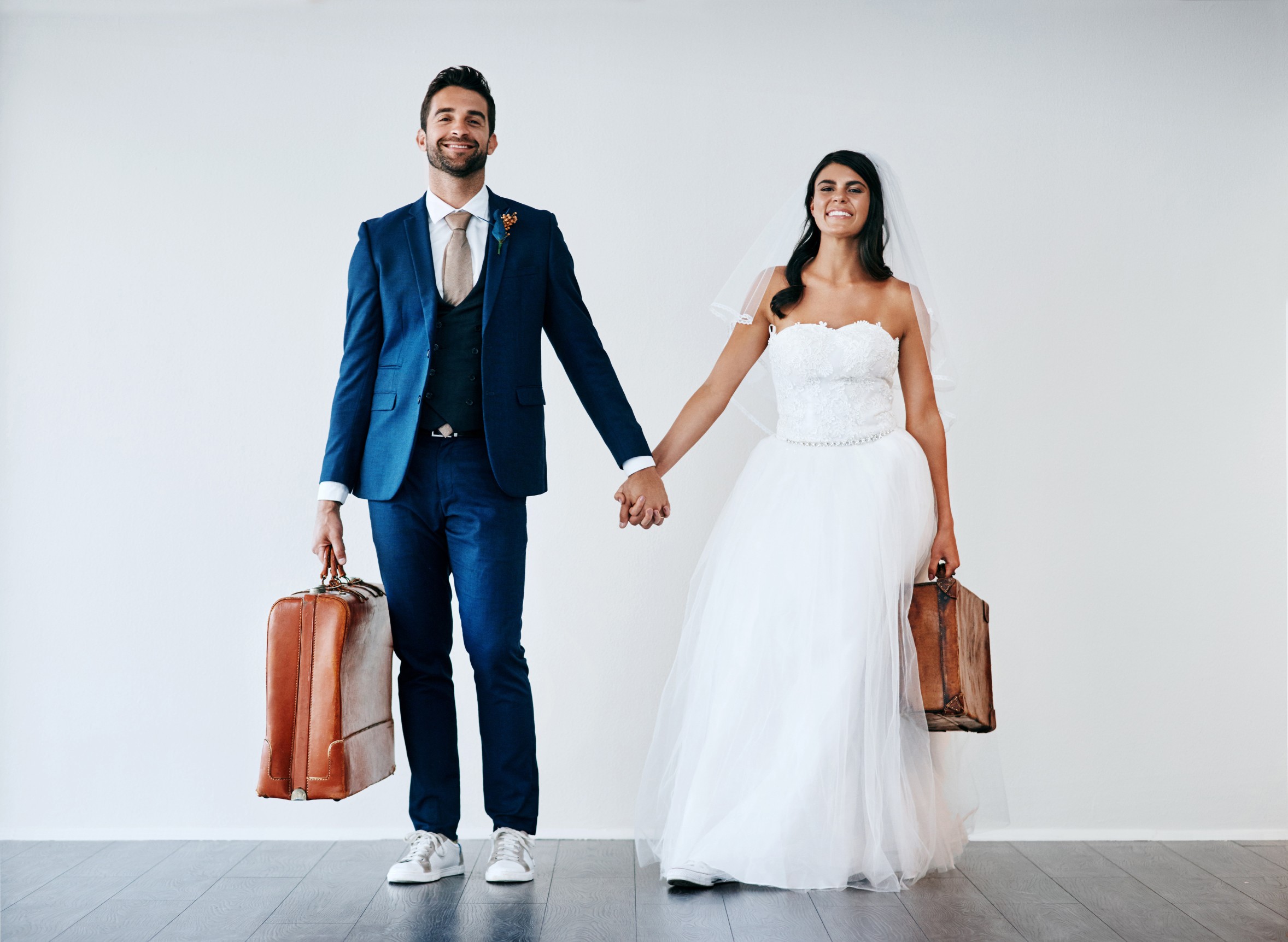 Bride and groom holding hands beside a suitcase, smiling in front of a white wall.