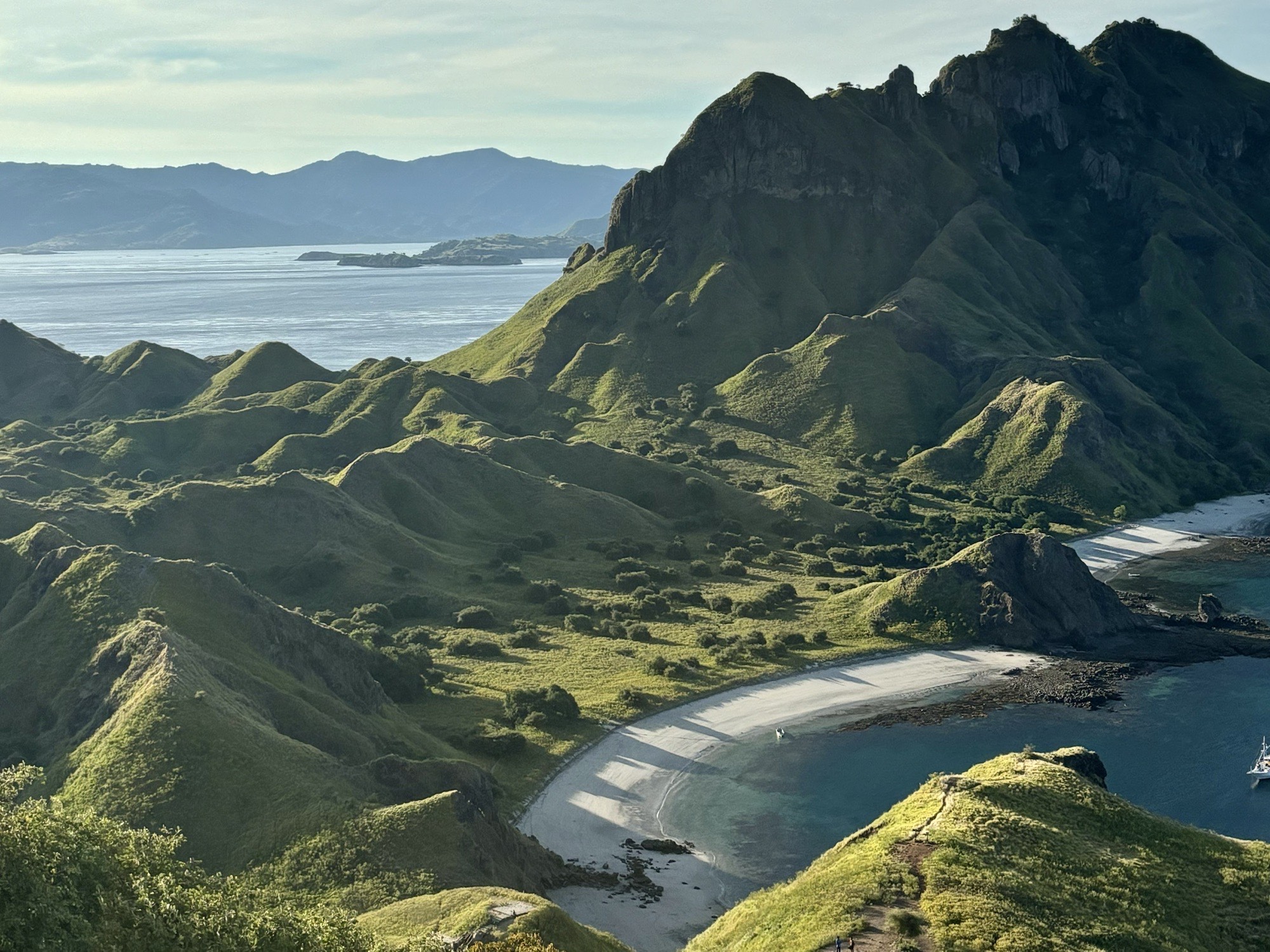 The image shows lush green hills rolling into crescent-shaped beaches and turquoise water, captured during a 4-day sailing adventure from Lombok to Labuan Bajo with Wanua Adventure.