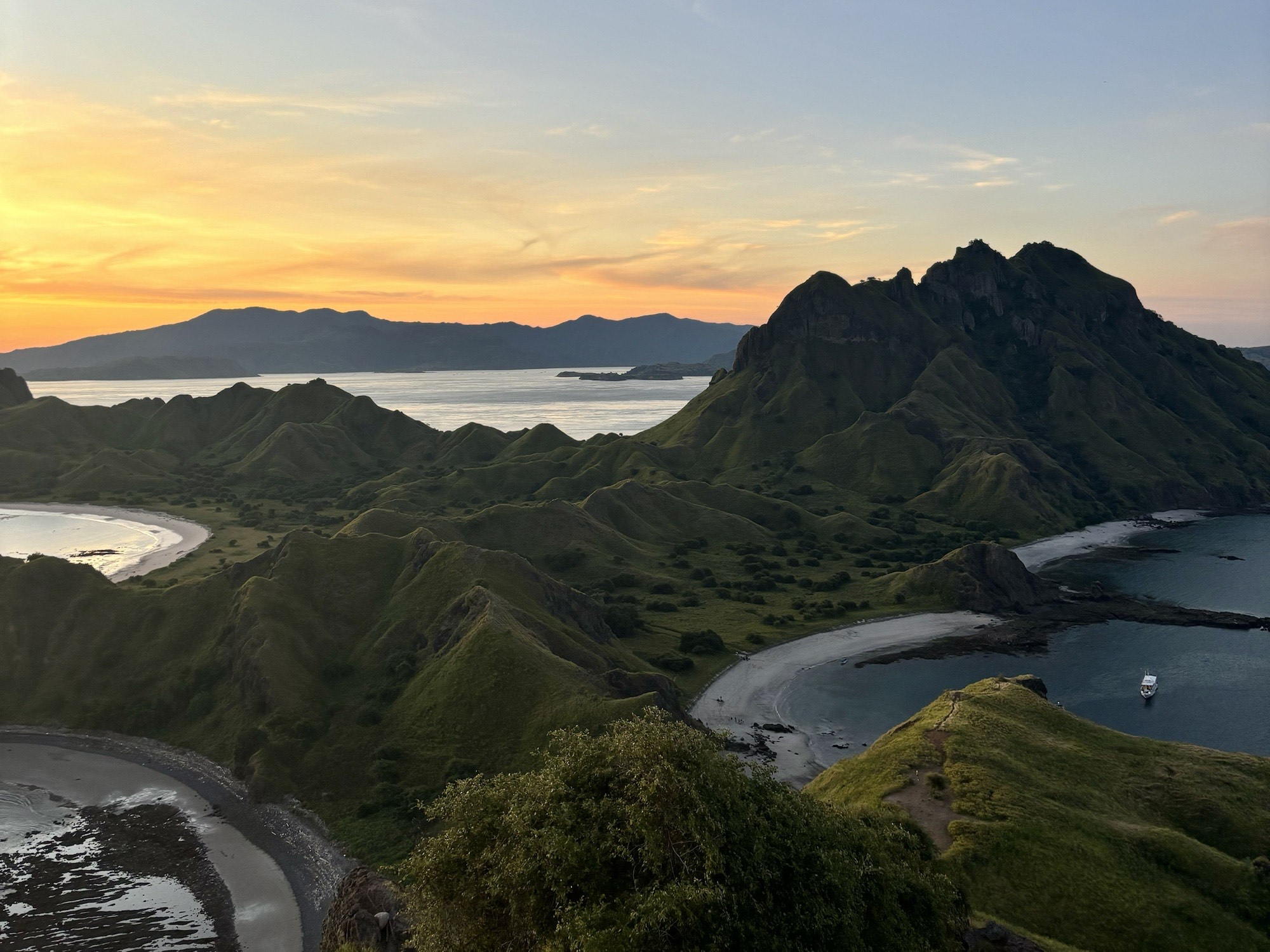 Dramatic aerial view of Padar Island in Komodo National Park, featuring rugged green hills, curved beaches, and vivid blue bays under the sunset.