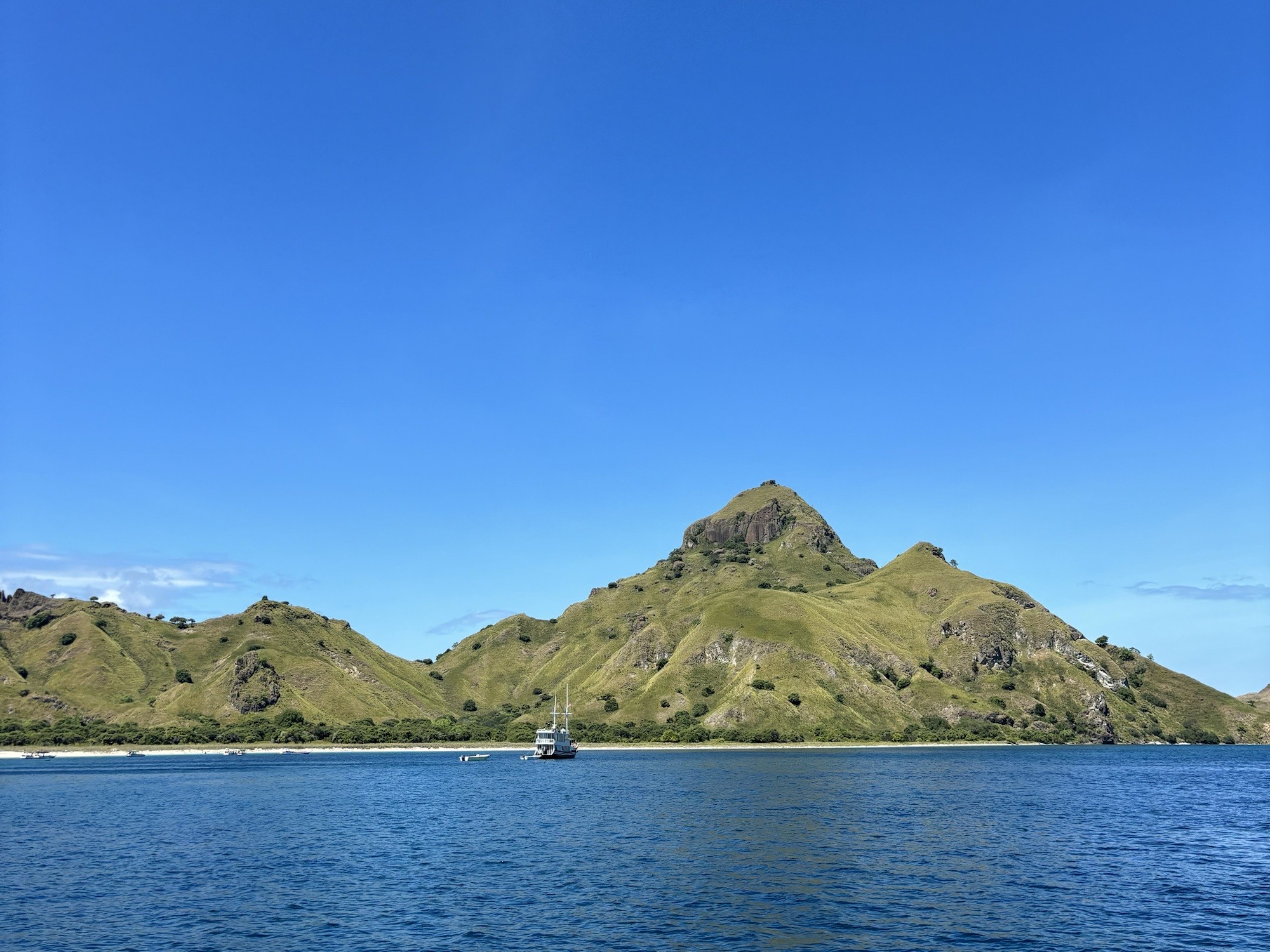 Scenic view of the Komodo Islands’ rolling green hills and rugged coastline, seen from a boat sailing through calm, turquoise waters.