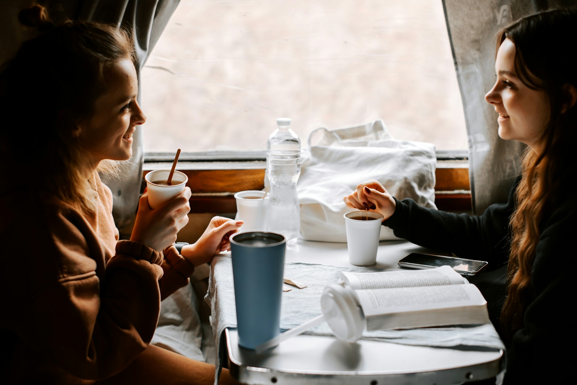 Two young women sip coffee and chat while on a train ride.