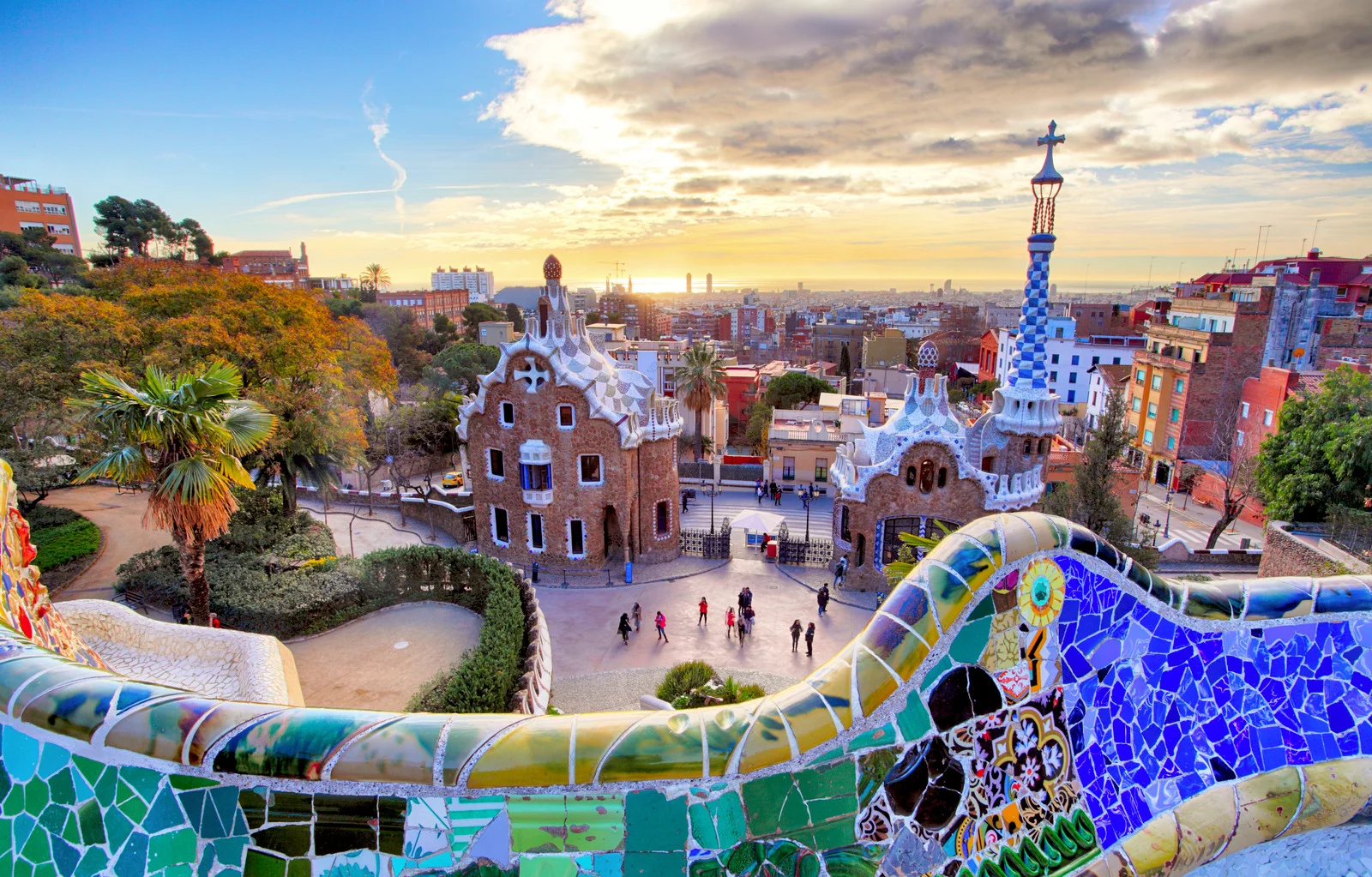 Colorful mosaic dragon fountain and curving tiled bench at Park Güell in Barcelona with panoramic city views.