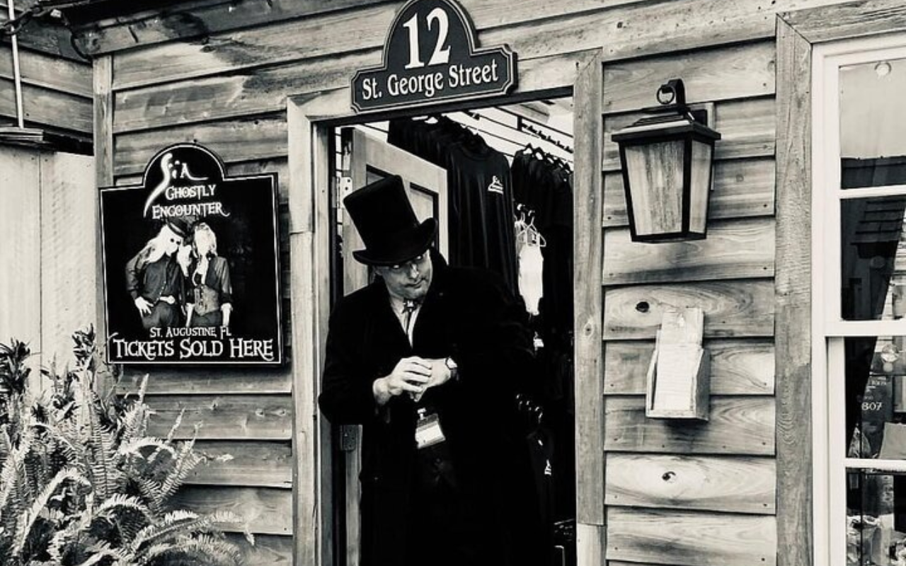 Man in a top hat and Victorian costume stands outside a ghost tour shop on St. George Street in St. Augustine, Florida, promoting haunted encounters.