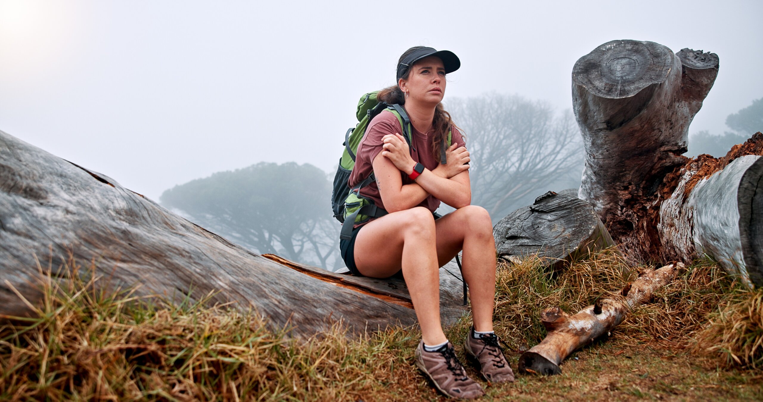 Girl, hiking and getting cold on mountain in winter, thinking and fatigue with weather change, fog and tourism. Person, woman and trekking on tree trunk, exhausted and tired on adventure in Argentina