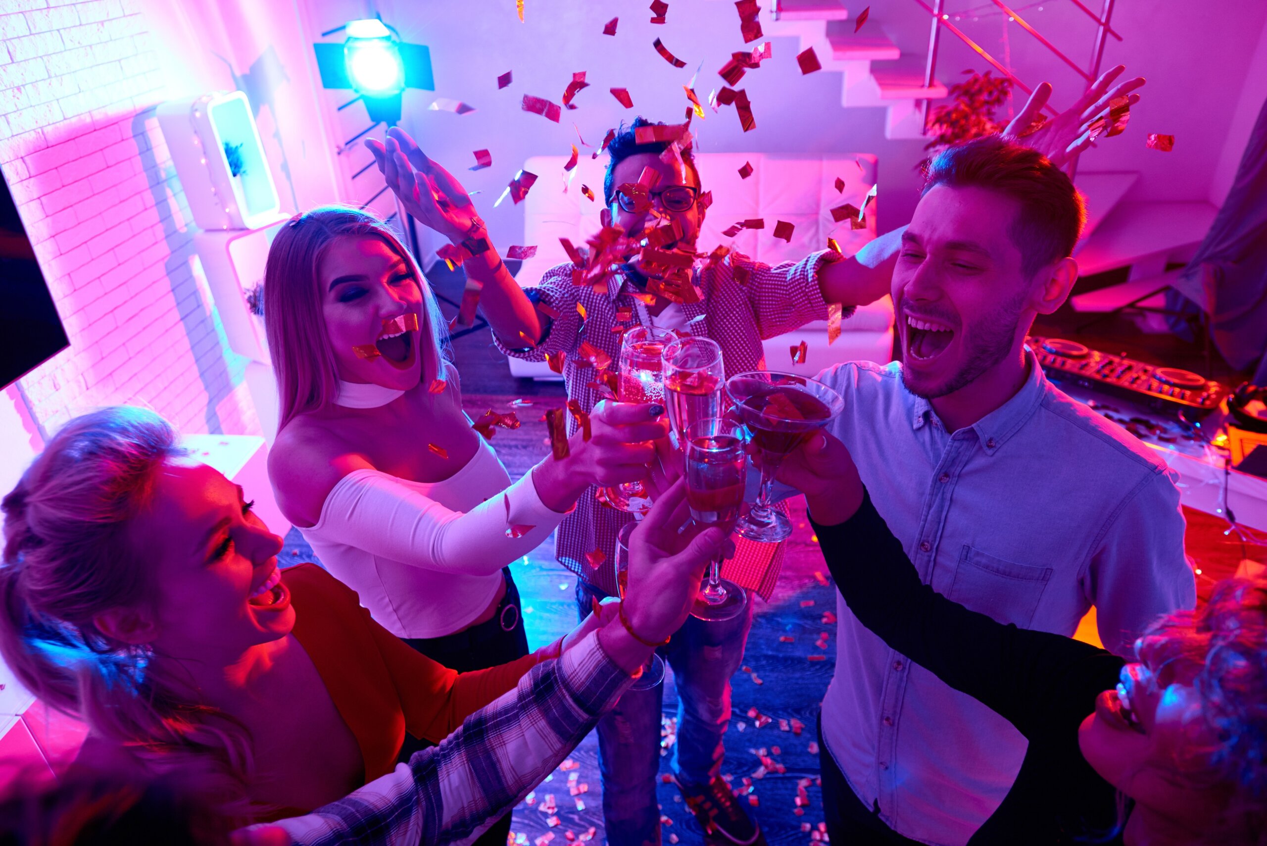 High angle view at group of happy friends enjoying holiday celebration at private house party raising glasses and toasting under bursts of confetti