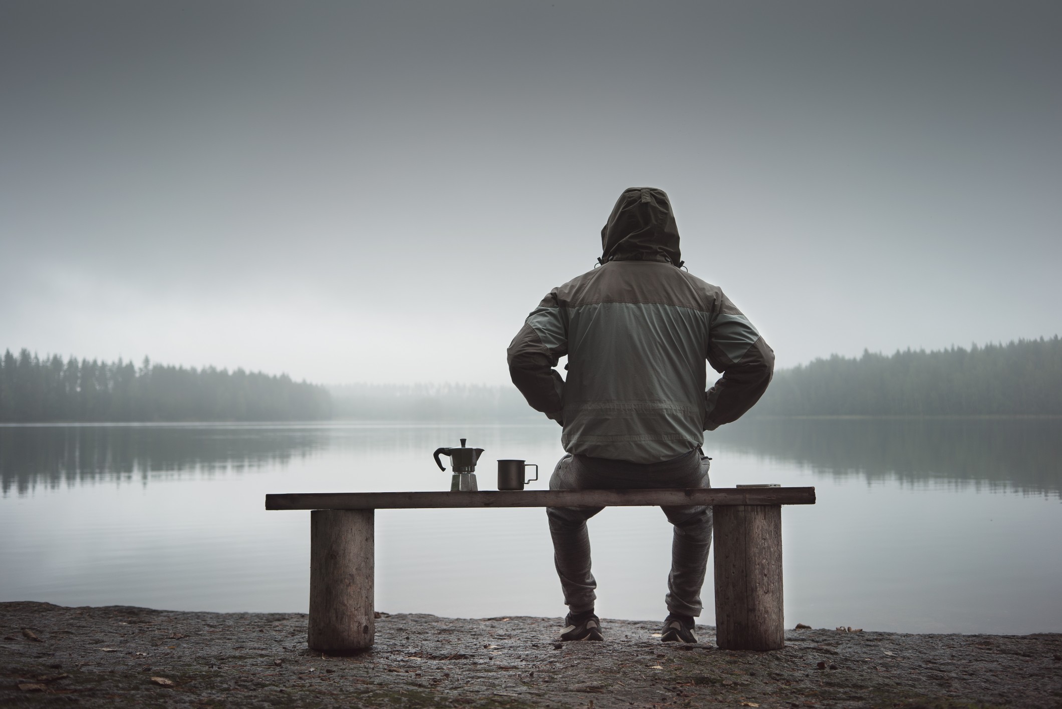 A man is sitting on a bench and looking at the lake. Back view. Also a coffee maker and a mug on the bench.
