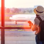 Traveler with backpack and hat at airport window preparing to travel abroad with travel insurance.