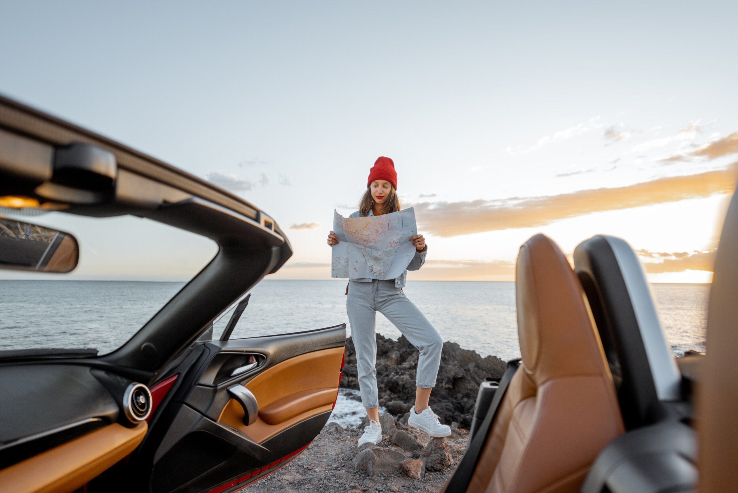 Woman enjoying beautiful view on the ocean, standing with map on the rocky coast during a sunset, wide view from the vehicle interior. Nature enjoyment and car travel concept