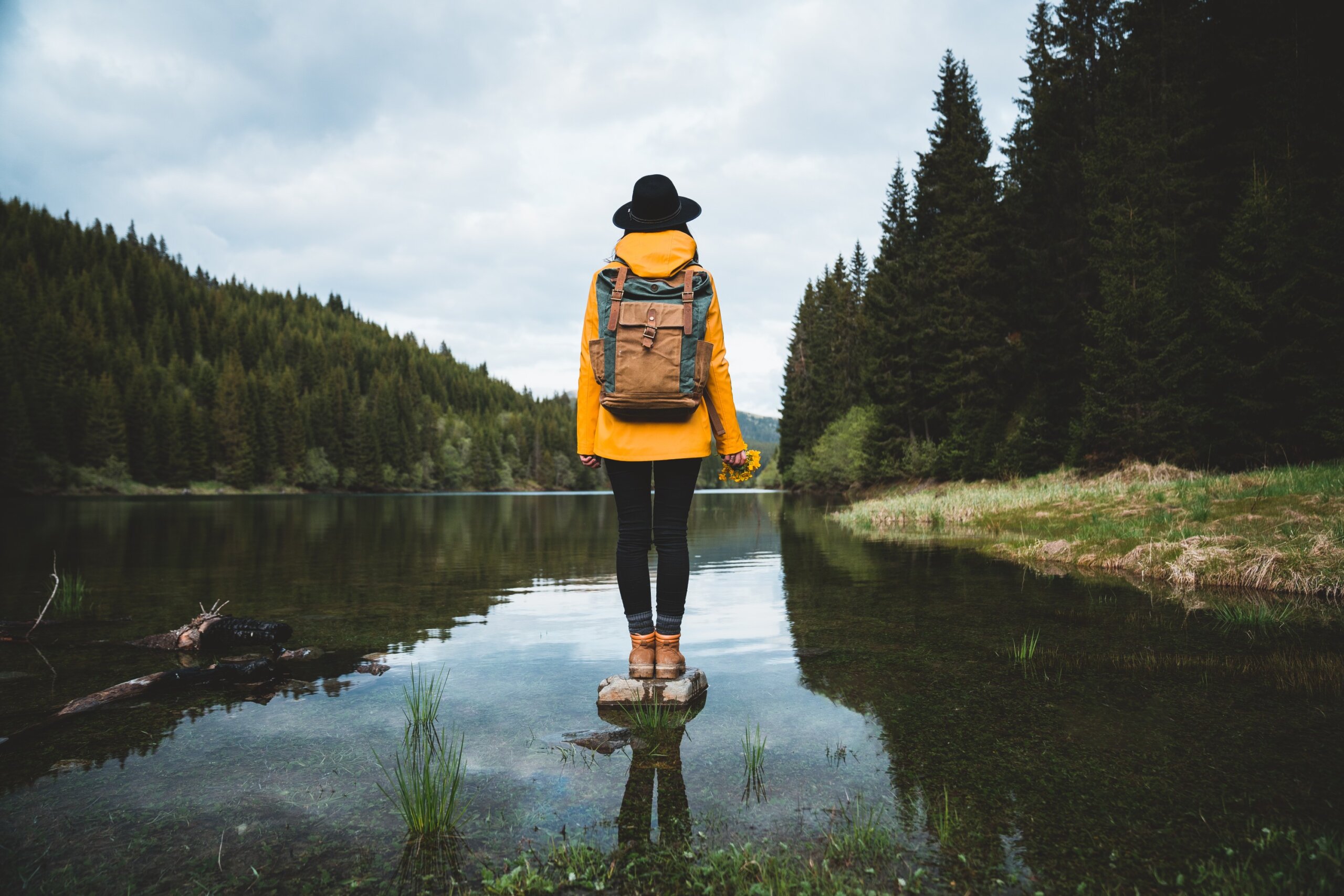 ack view of stylish hipster woman holding flowers,wearing vintage backpack, hat and yellow jacket looking at mountain view while relaxing in nature. Travel and wanderlust concept.