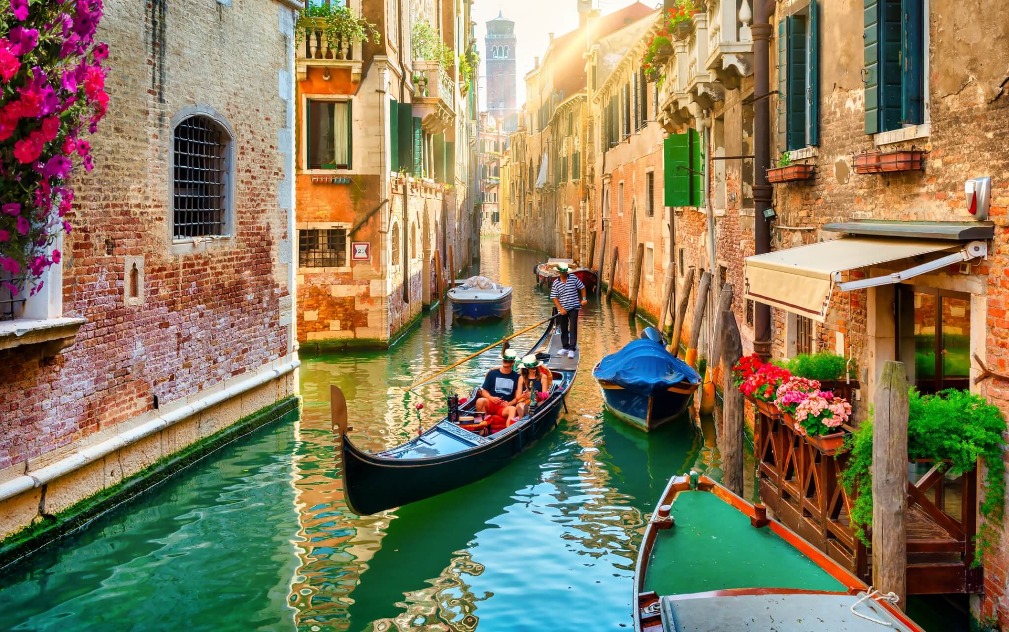 Tourists riding a gondola through a scenic canal in Venice, Italy, with historic buildings and flowers along the water.