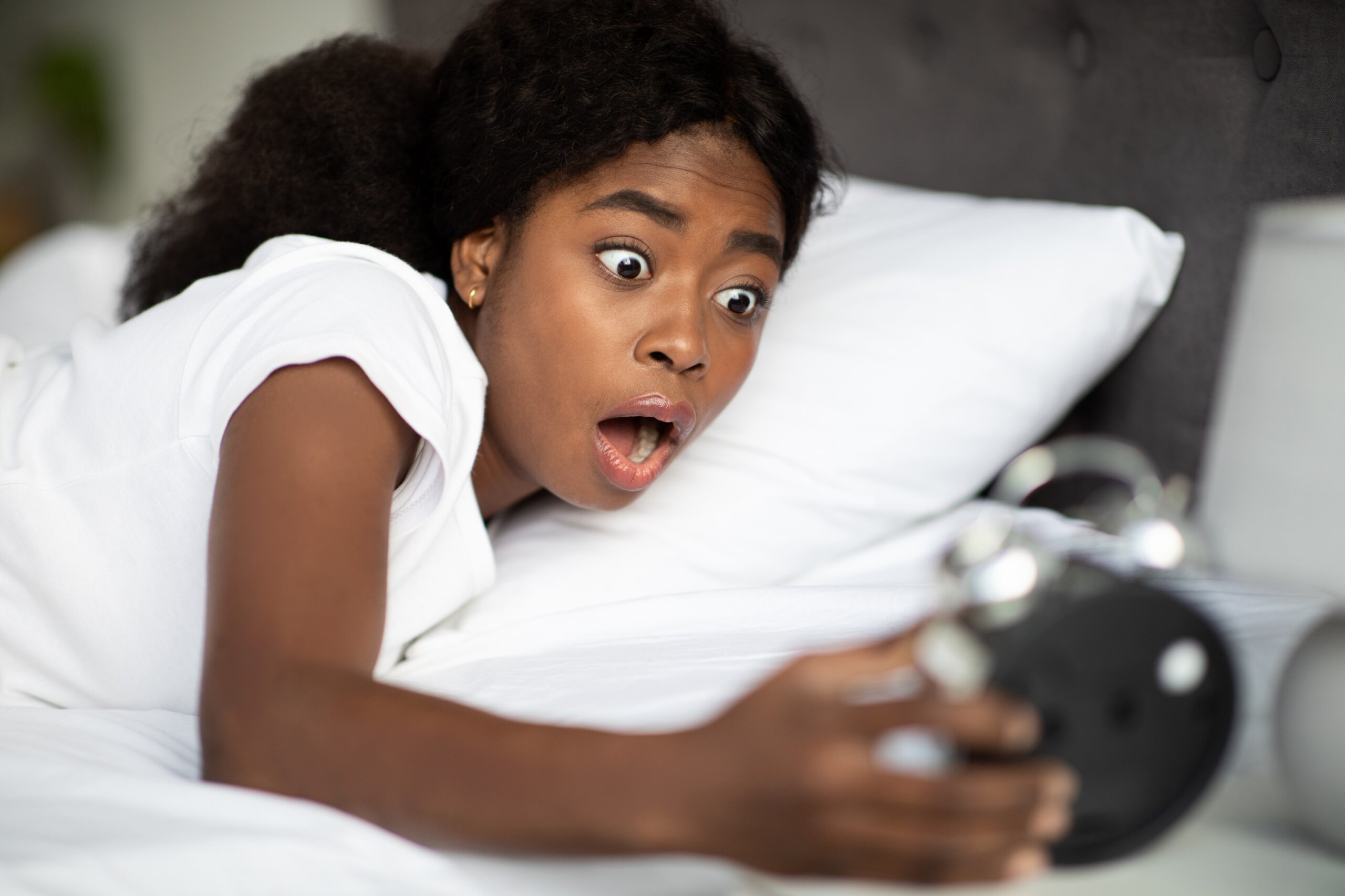Woman hold big black clock on bed in bedroom at home. She shock after wake up too late to catch the bus for her group tour.