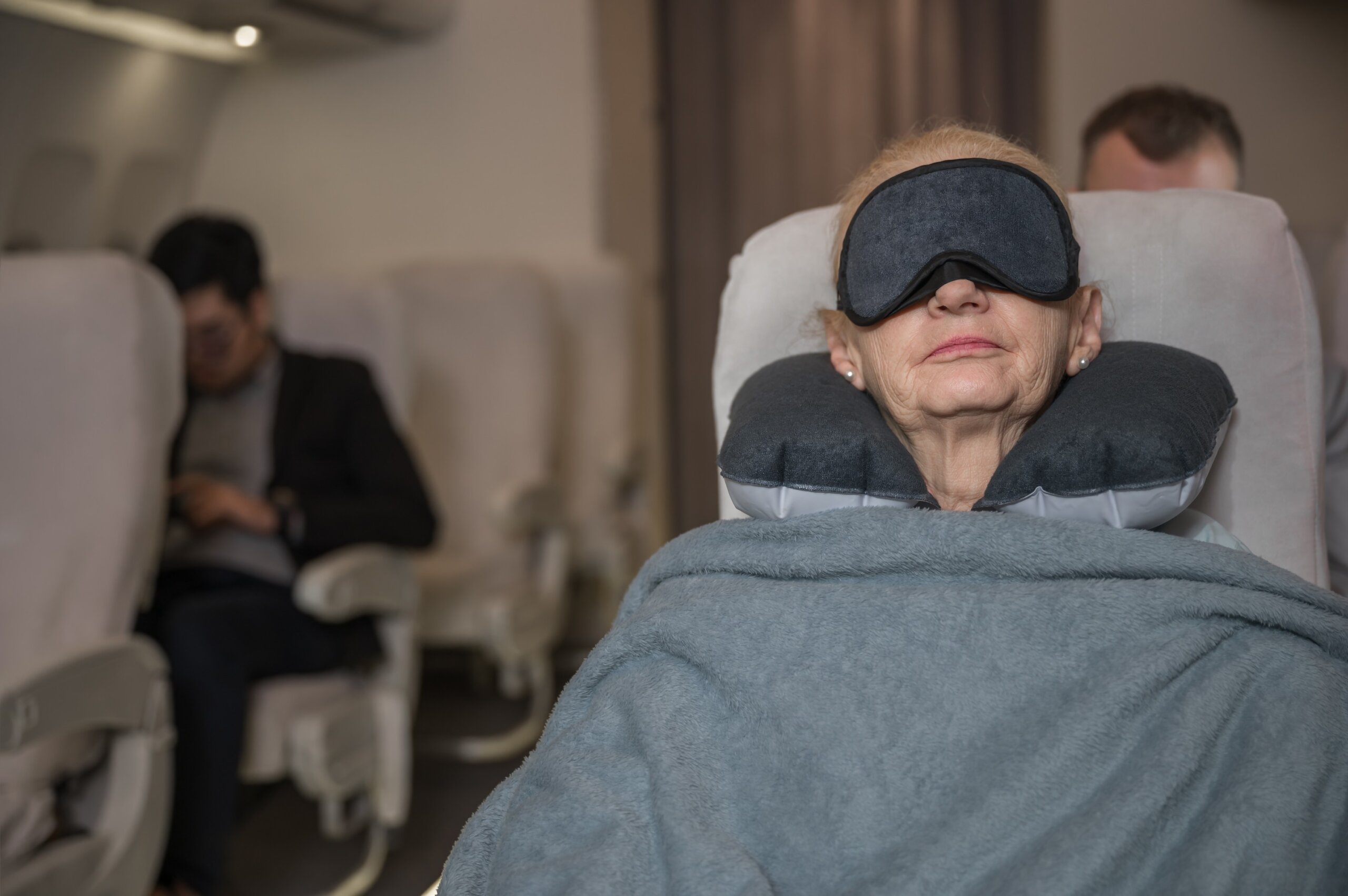Happy elderly woman passenger use blindfold mask and neck pillow with a comfortable blanket while sleeping on airplane seat in flight.