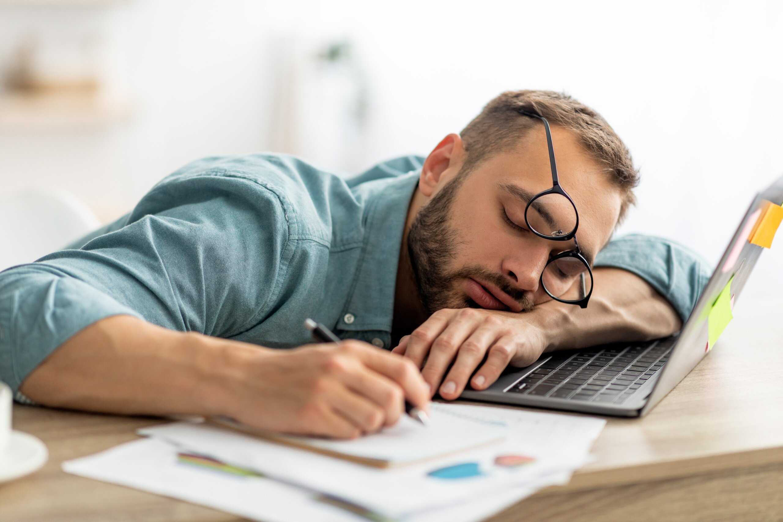 Exhausted millennial man sleeping on his office desk, next to laptop and documents