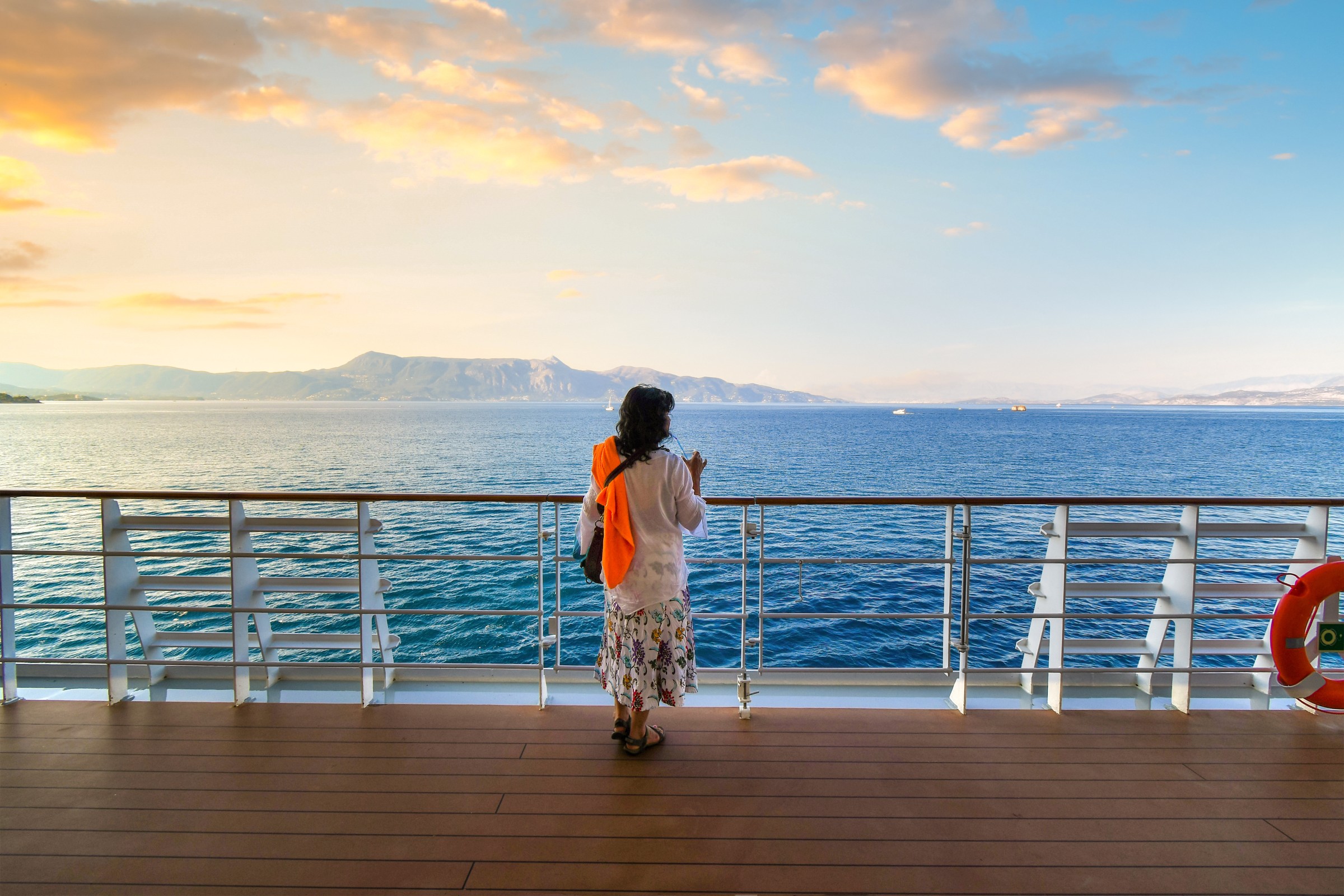 A woman stands on the deck of a cruise ship at sunset while it cruises the Mediterranean Sea surrounded by Greek Islands.
