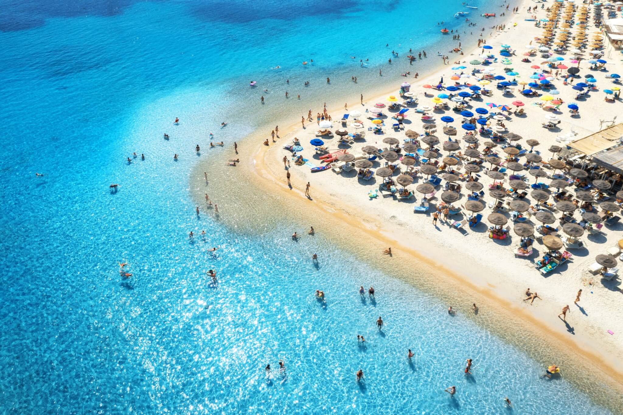 Aerial view of colorful umbrellas on sandy beach, people in blue sea at sunset in summer. Tuerredda Beach, Sardinia, Italy. Tropical landscape with turquoise water. Travel and vacation.